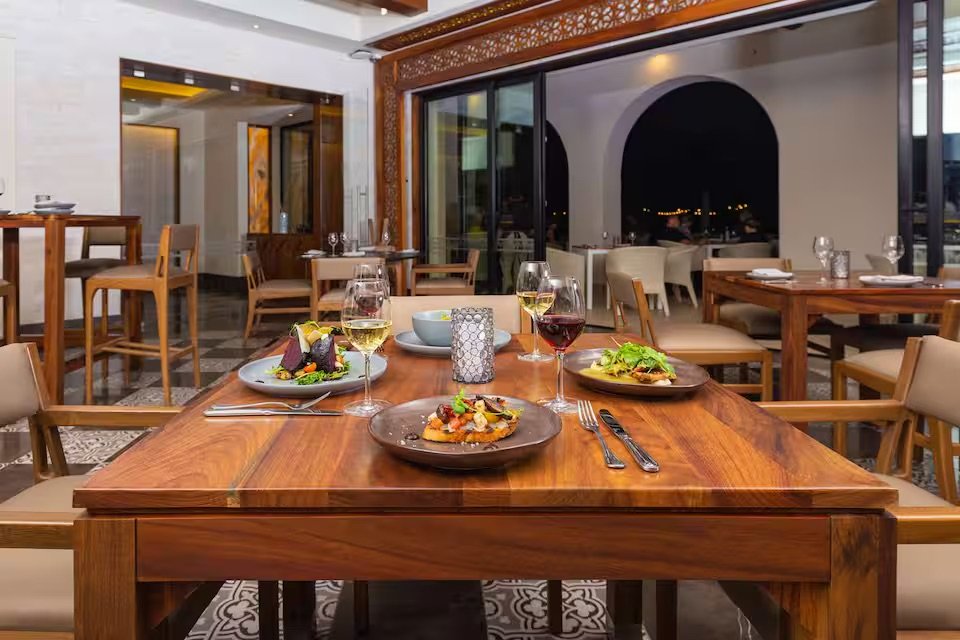A dining table in a restaurant set with plates of food, glasses of white and red wine, and cutlery, with empty chairs and a view of the night outside through large windows.