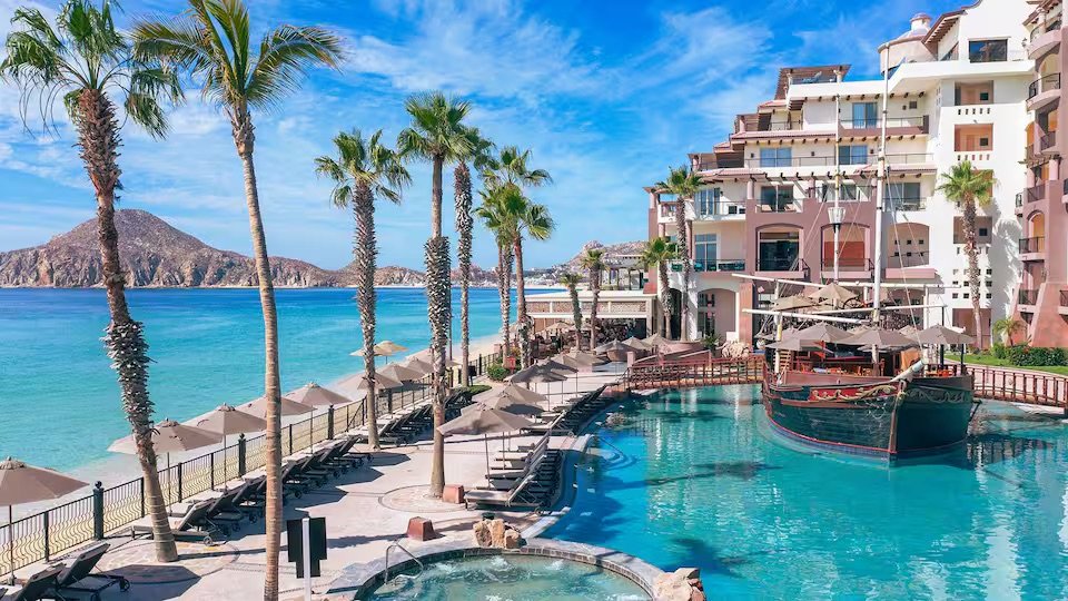 Luxury poolside scene at a beach resort featuring palm trees, lounge chairs with umbrellas, a swimming pool with a boat docked on the edge, white multi-story resort buildings, a sandy beach, and distant hills under a blue sky with some clouds.