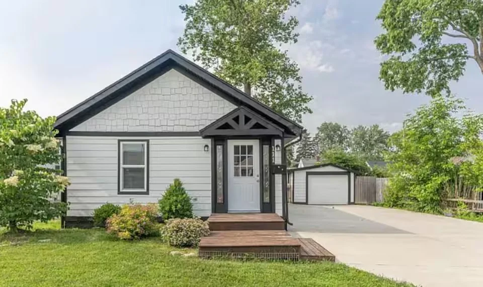 Small white house with a gabled roof, front porch with steps, and a concrete driveway leading to a garage, surrounded by green trees and lawn.