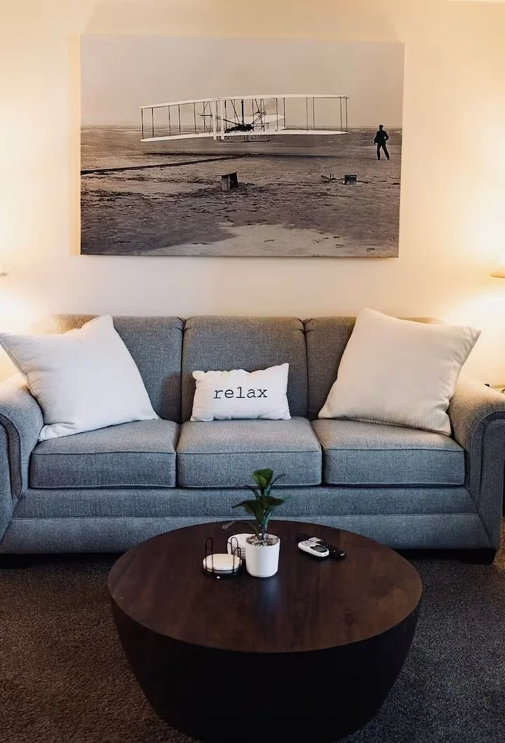 A living room with a gray sofa, white pillows, and a small white pillow with the word "relax" on it. Above the sofa is a black-and-white picture of the Wright brothers' airplane on a beach. A wooden coffee table with a small plant, a remote control, and a couple of candles is in front of the sofa.