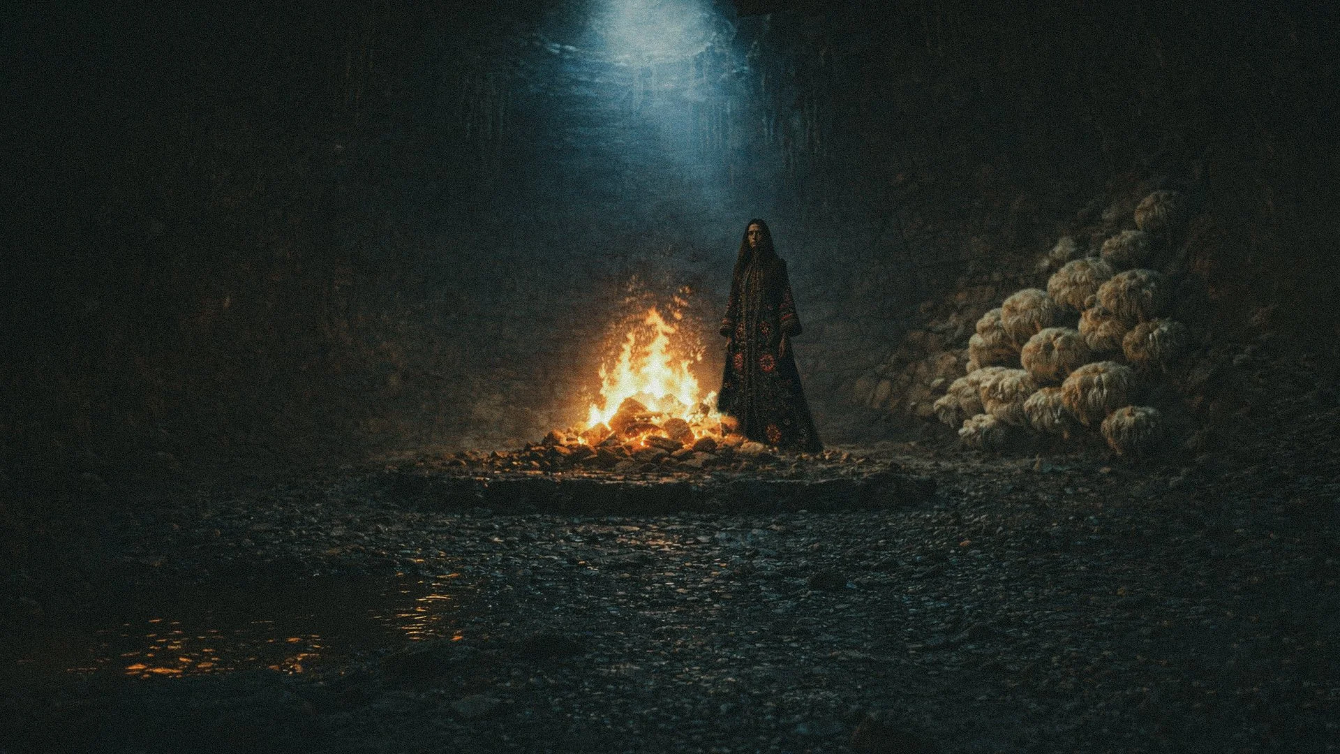 A woman in a long, dark, patterned dress standing in a dim, rocky underground tunnel with a fire and pile of rocks behind her and a large group of sheep lined up on the right side.