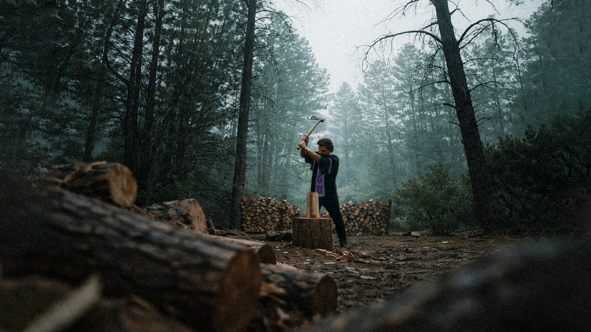 A person chopping wood with an axe in a dense forest surrounded by logs and cut wood piles.