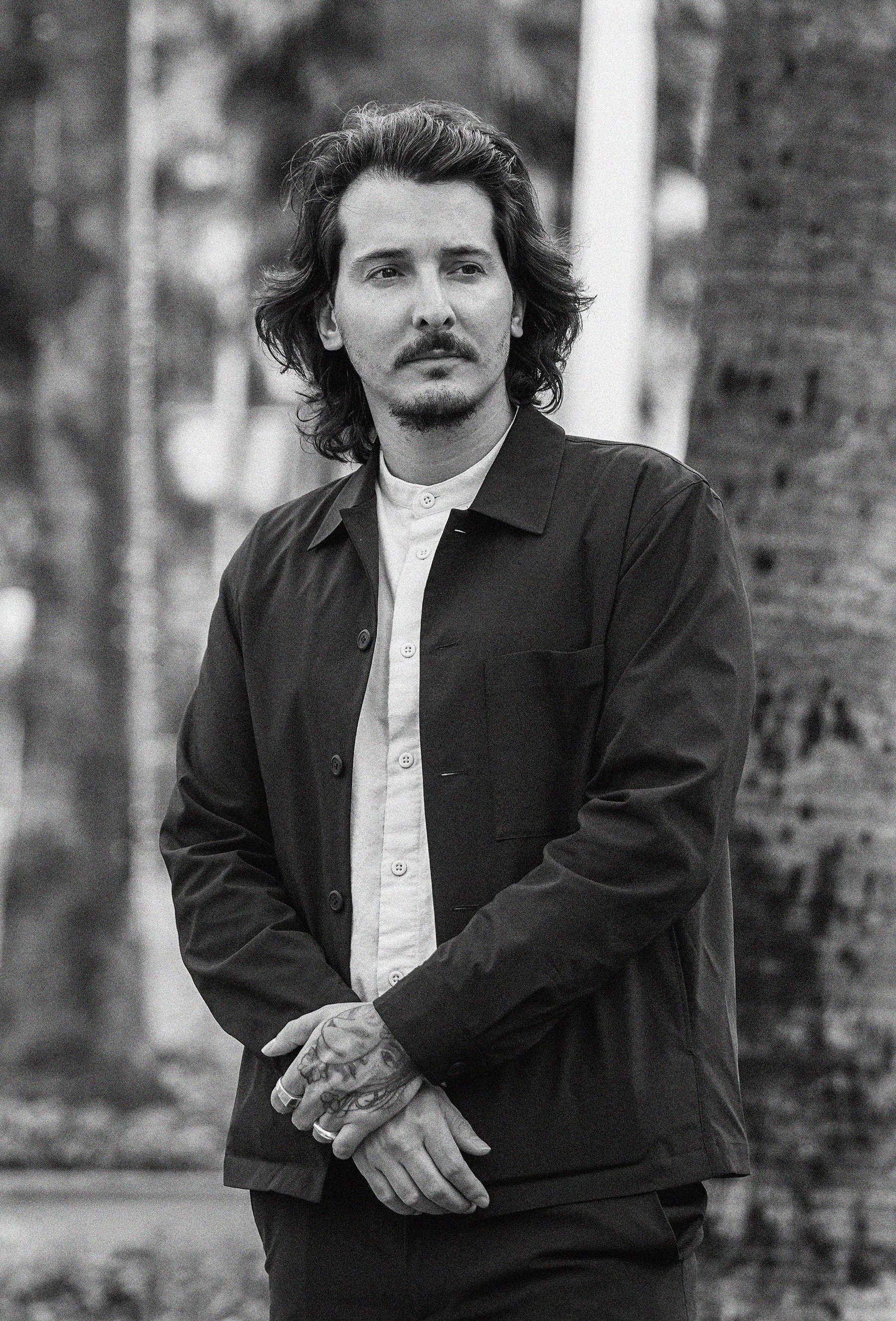 A black and white photo of a man with long hair and a mustache, wearing a button-up shirt and a jacket, standing outdoors with trees and a brick wall in the background.
