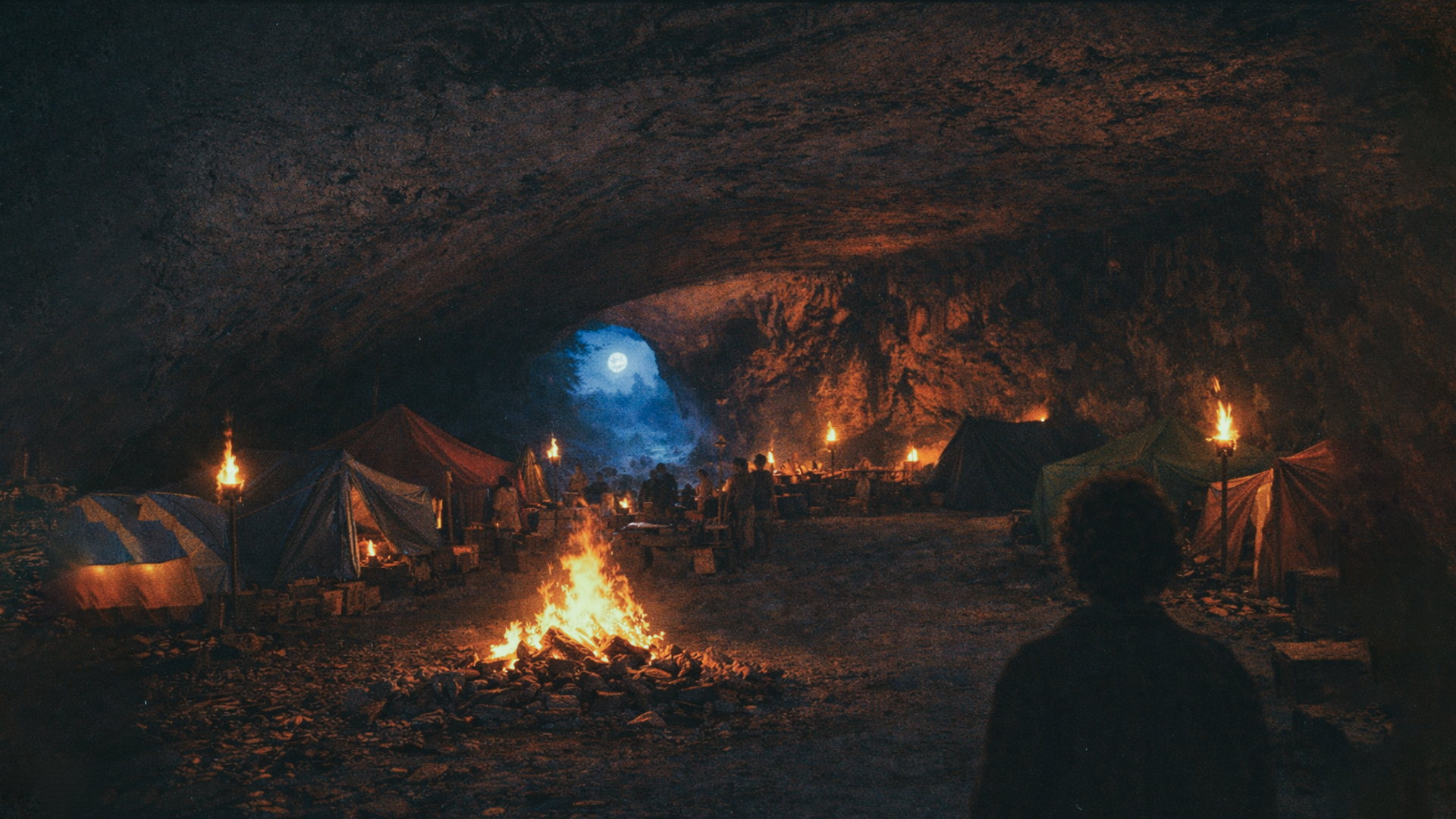 Underground campsite with tents and a campfire, illuminated by torches, under a large cave ceiling, with a person in the foreground and the full moon visible through an opening in the cave.