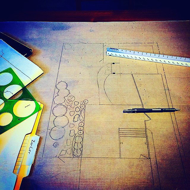 Sketch of a landscape design with trees, rocks, and pathways on a wooden table, with drawing tools including a pen, a ruler, and a drafting stencil.