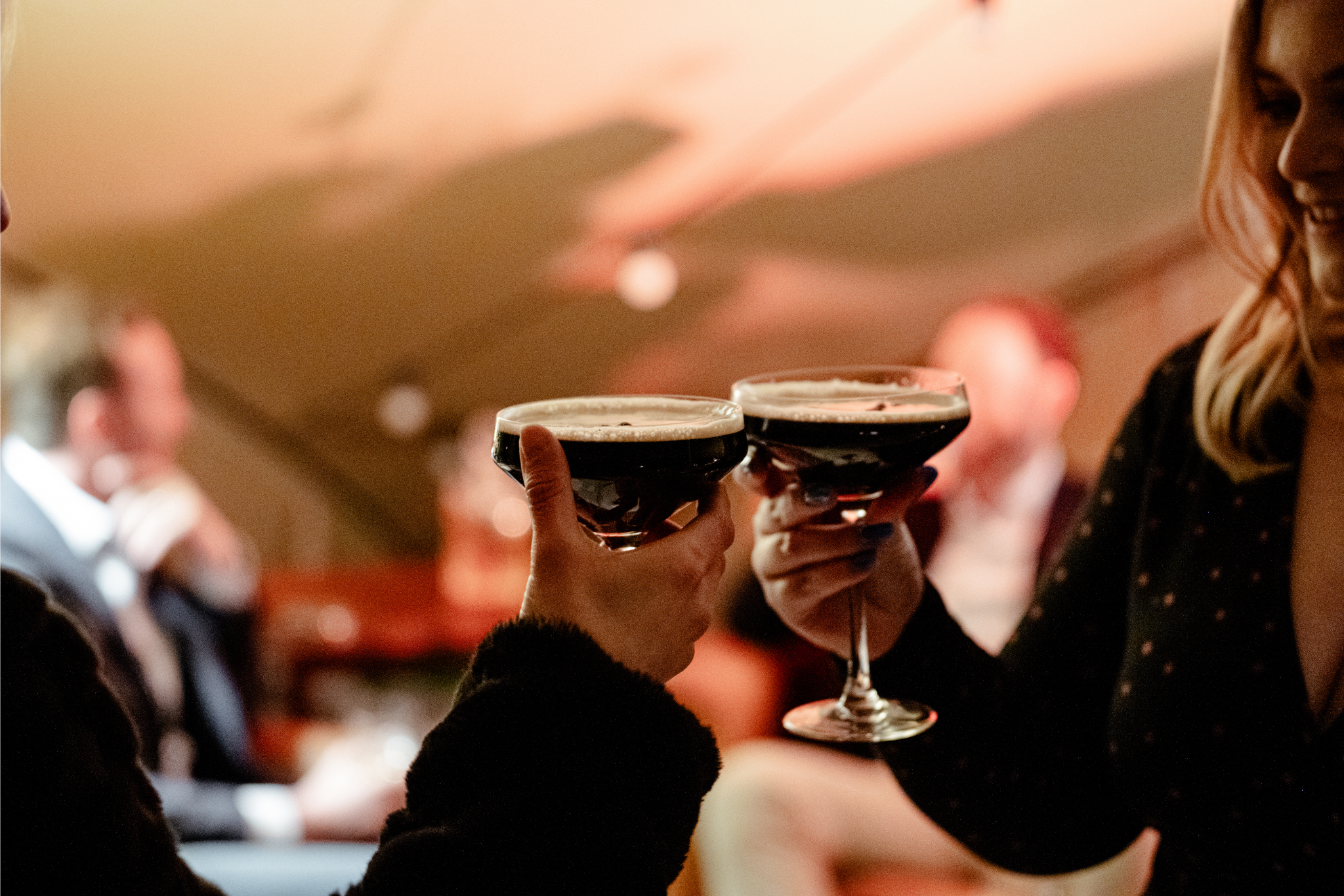 Two women are raising glasses of dark beer for a toast at a social event.