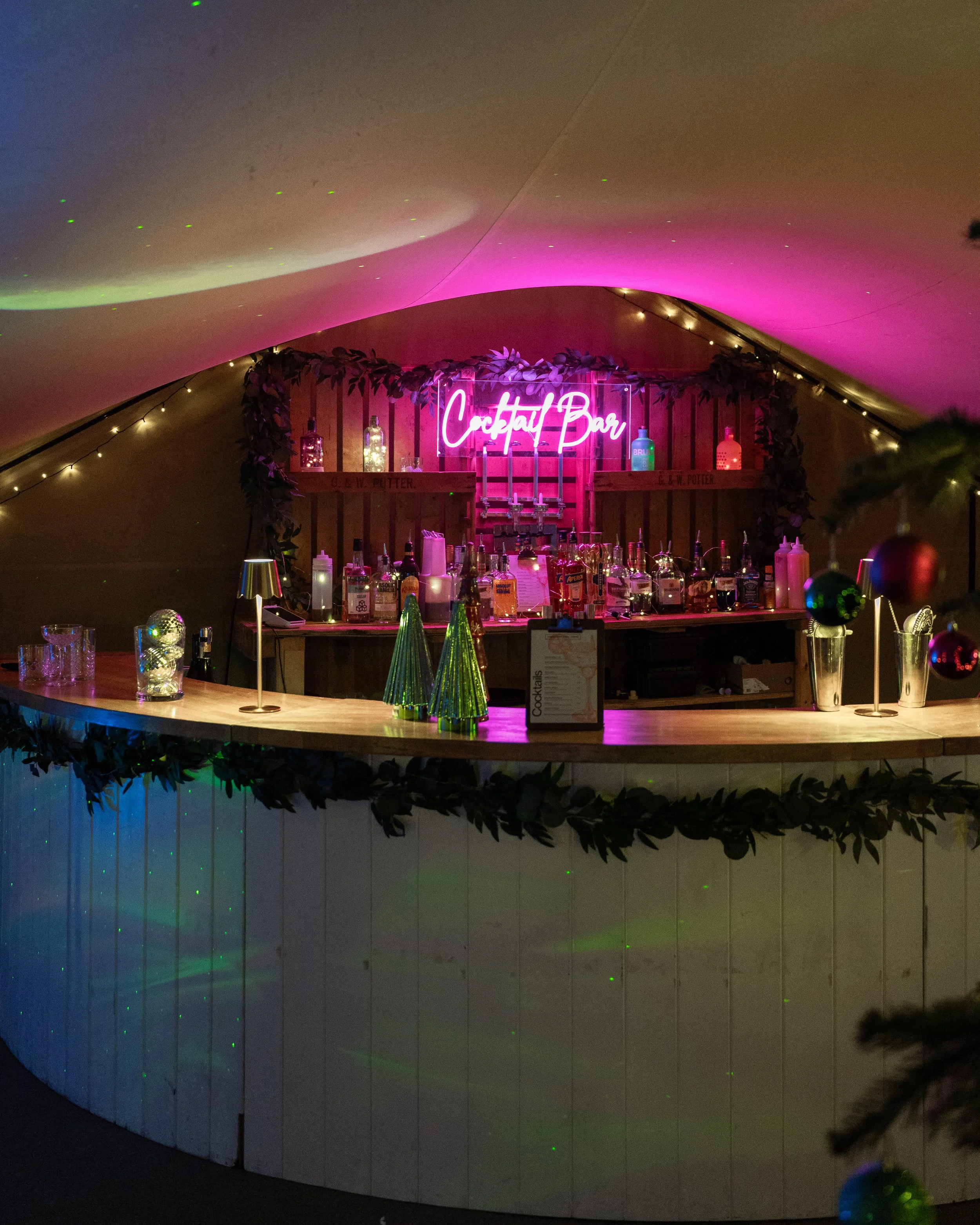 A decorated bar area with a neon pink sign that says 'Cocktail Bar,' illuminated bottles behind the counter, and holiday decorations including small Christmas trees and ornaments.