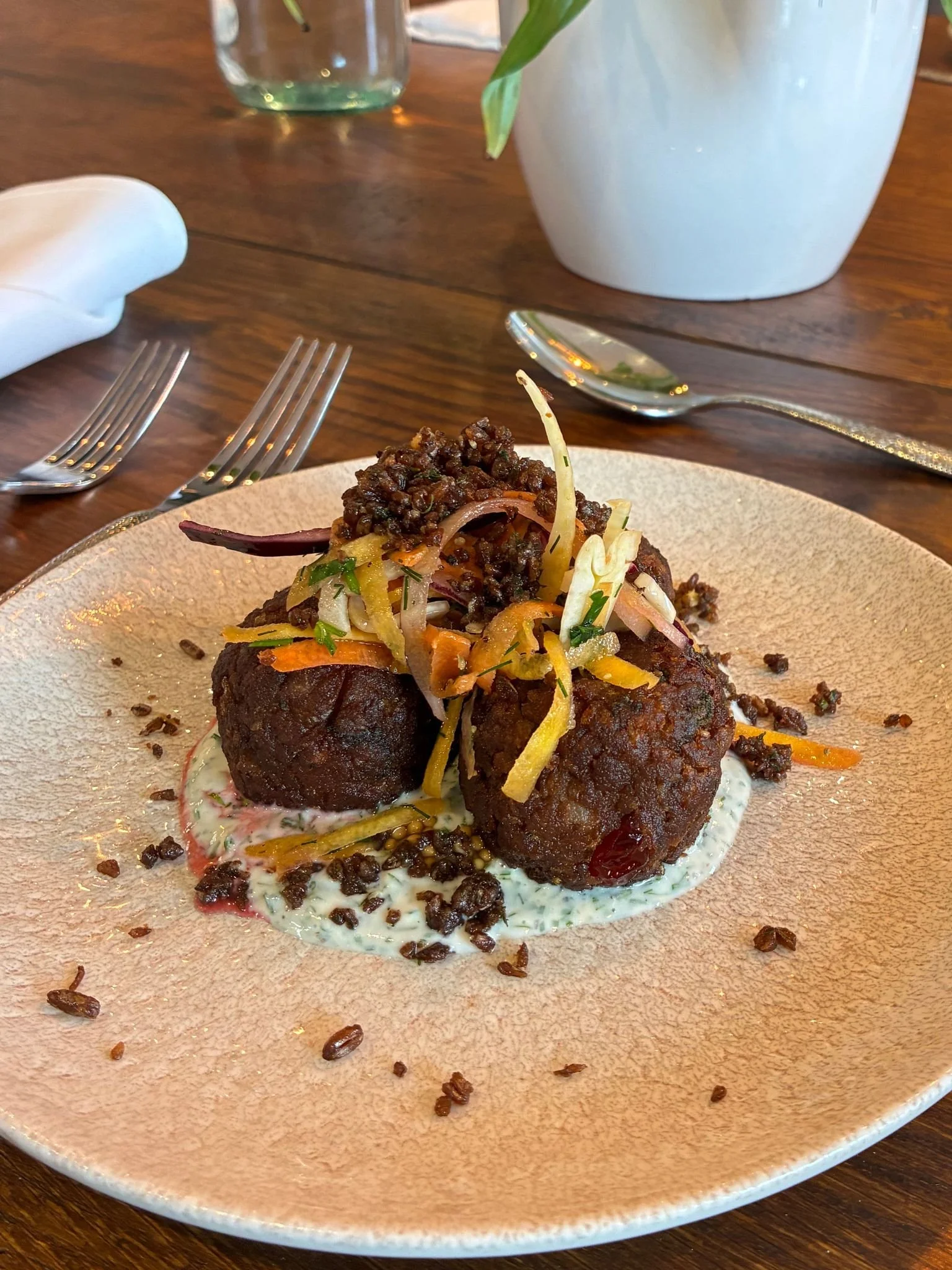 A plate with two large meatloaf or potato croquettes topped with vegetables on a textured beige plate. Plate is on a wooden table with silverware, a white napkin, a glass of water, and a vase with green leaves in the background.