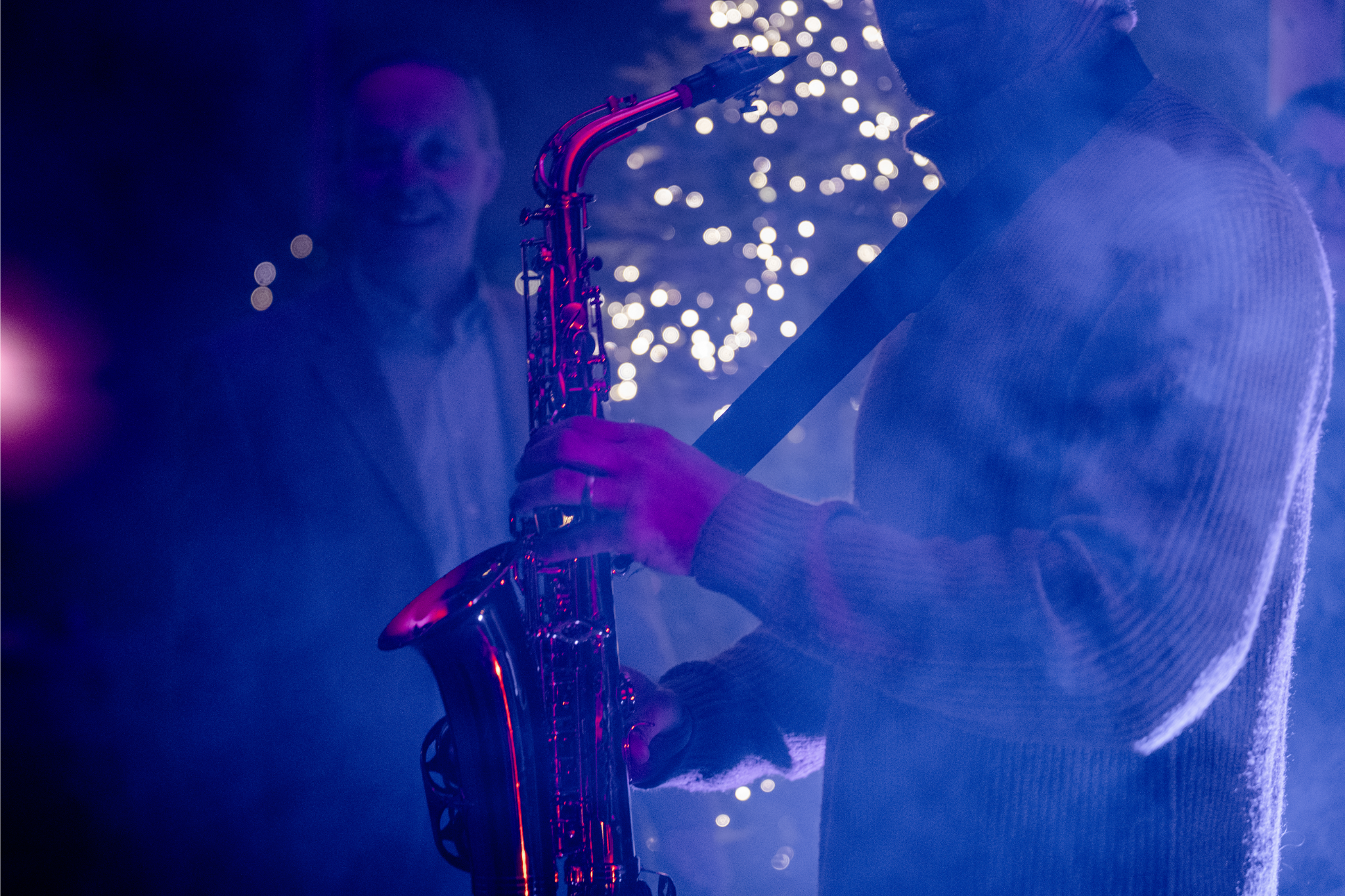 A person playing the saxophone in a dimly lit setting with purple and blue lighting and blurred bokeh lights in the background.