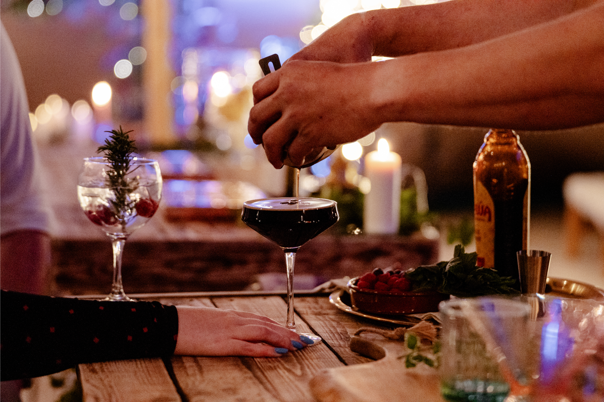 A person pouring dark red wine into a martini glass at a cozy, warmly lit bar or restaurant, with holiday decorations and candles in the background.