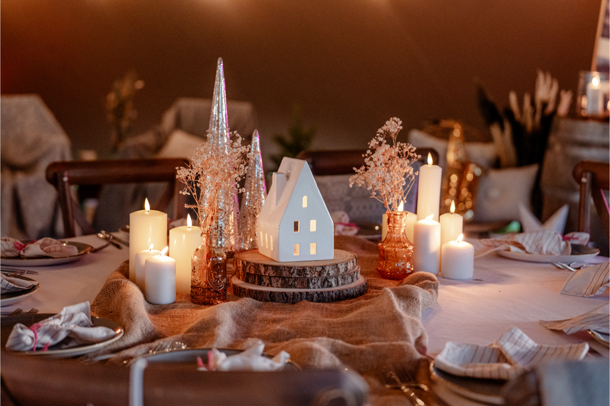 A festive table centerpiece with candles, dried flowers, and decorative trees arranged on a table with a burlap runner, set for a holiday or Christmas celebration.