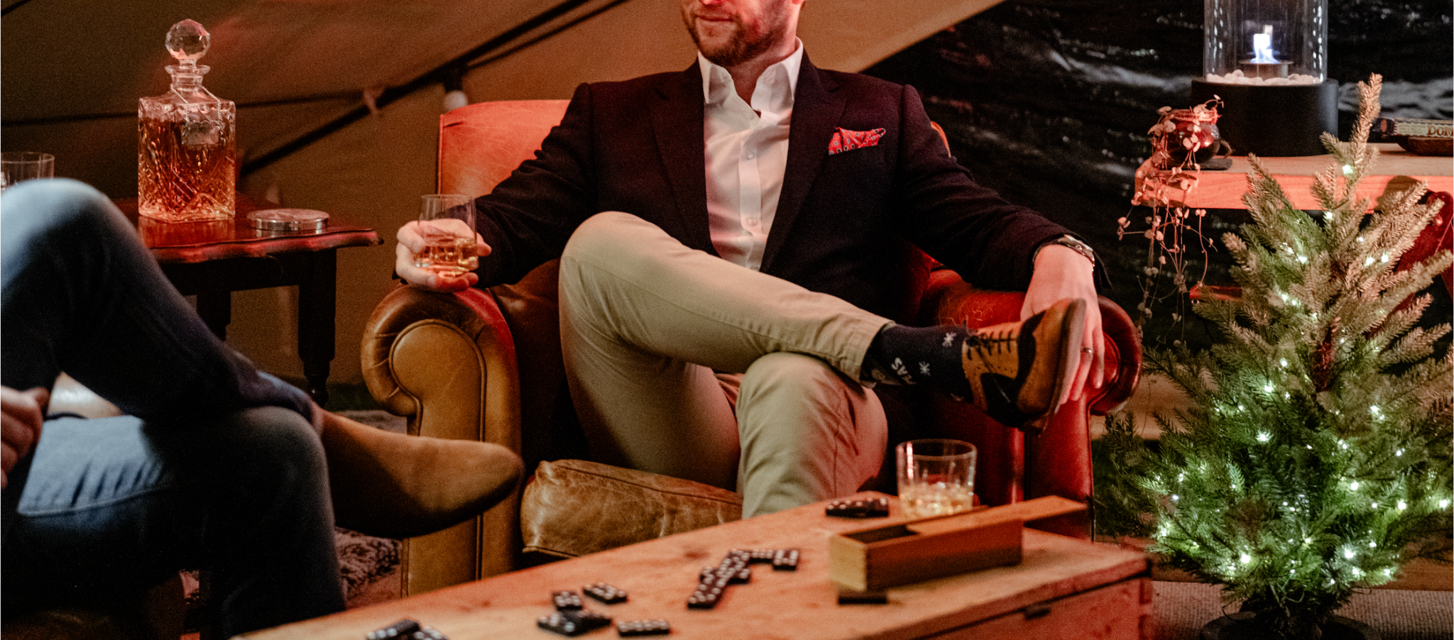 A man in a dark blazer, white shirt, tan pants, and colorful socks is sitting relaxed on a leather armchair, holding a glass of amber liquor, with a Christmas tree decorated with lights nearby. A wooden table with drinks and decorations is in front o
