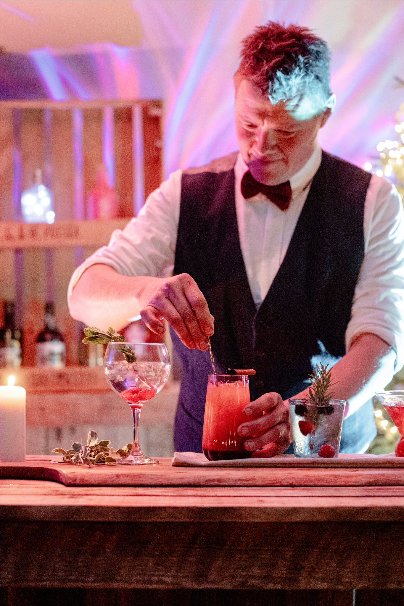 A bartender wearing a white shirt, black vest, and bow tie preparing colorful cocktails behind a wooden bar with candles and festive decorations.