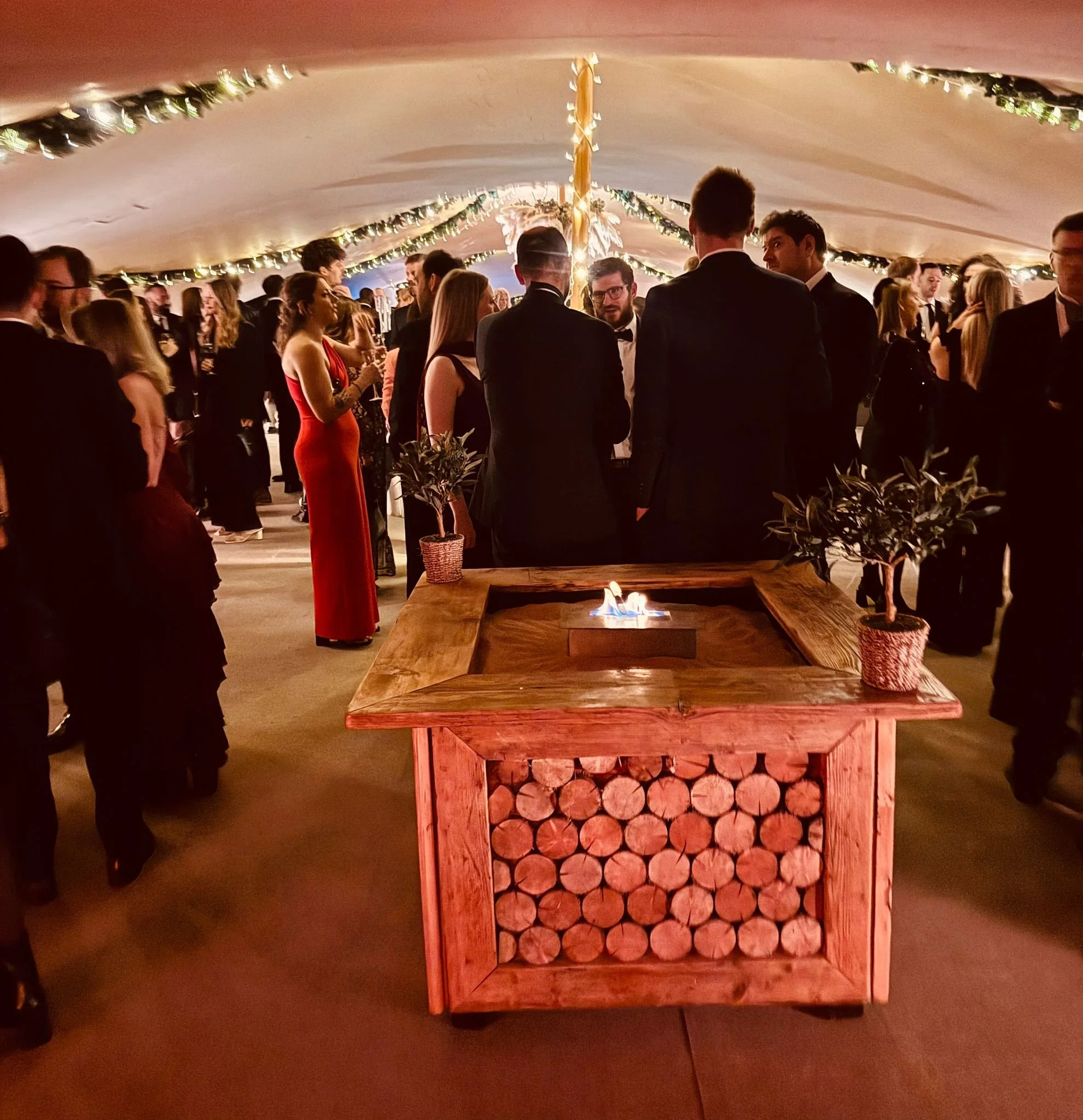 A wedding reception with guests in formal attire under a decorated tent, featuring a wooden chafing dish with logs and a small flame in the foreground, and string lights and greenery hanging from the ceiling.