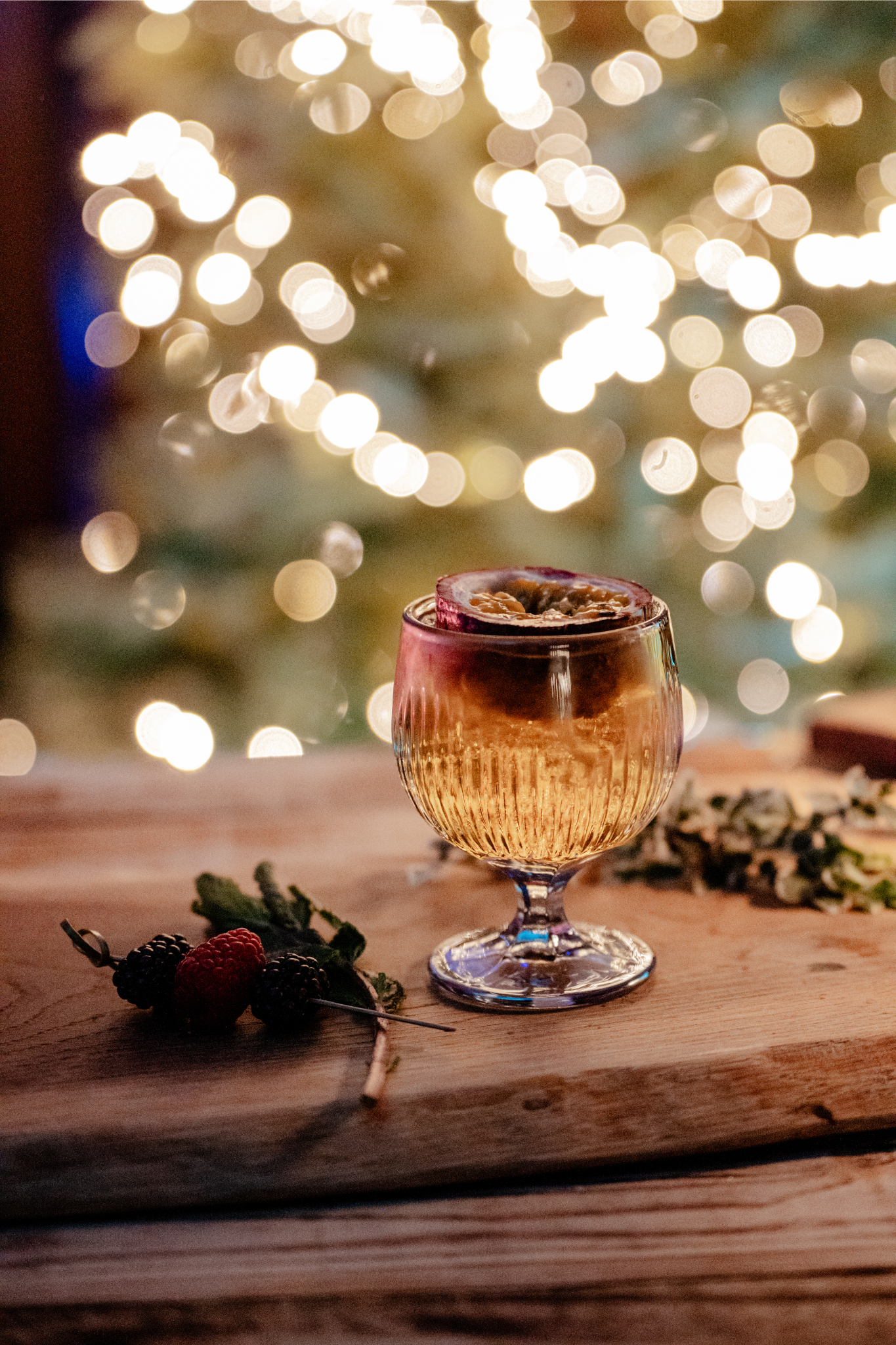A cocktail with a dried citrus slice garnish on a wooden surface, with berries and herbs nearby, and blurred festive string lights in the background.