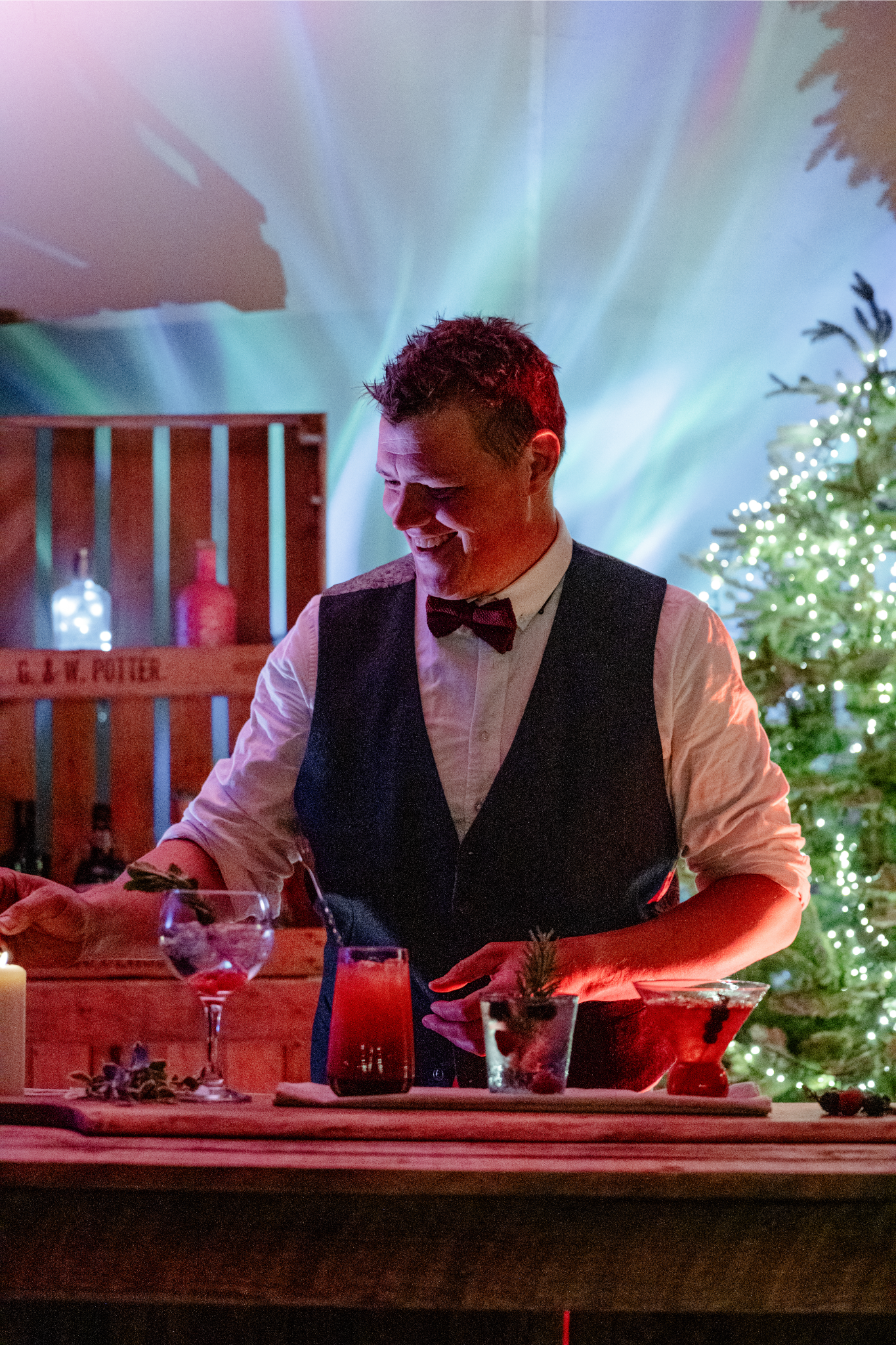 A man dressed as a bartender or host, wearing a vest and bow tie, smiling while preparing drinks at a festive holiday party with Christmas decorations, including a lit Christmas tree and assorted bottles in the background.
