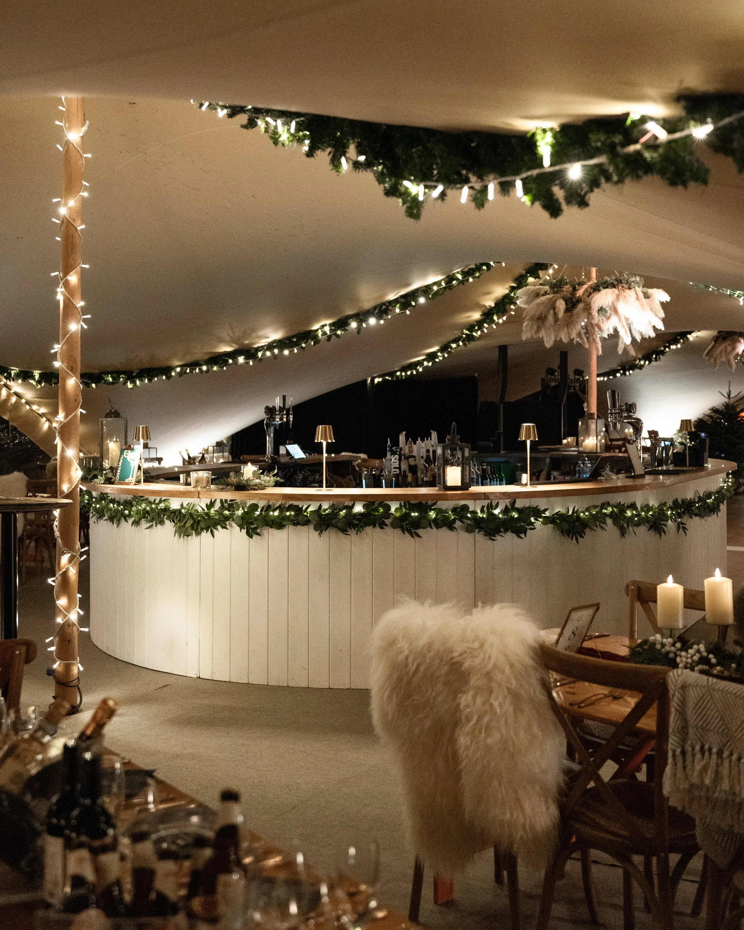 Decorated bar area with string lights, garlands, and candles, featuring a cozy and festive atmosphere, with chairs and tables nearby.