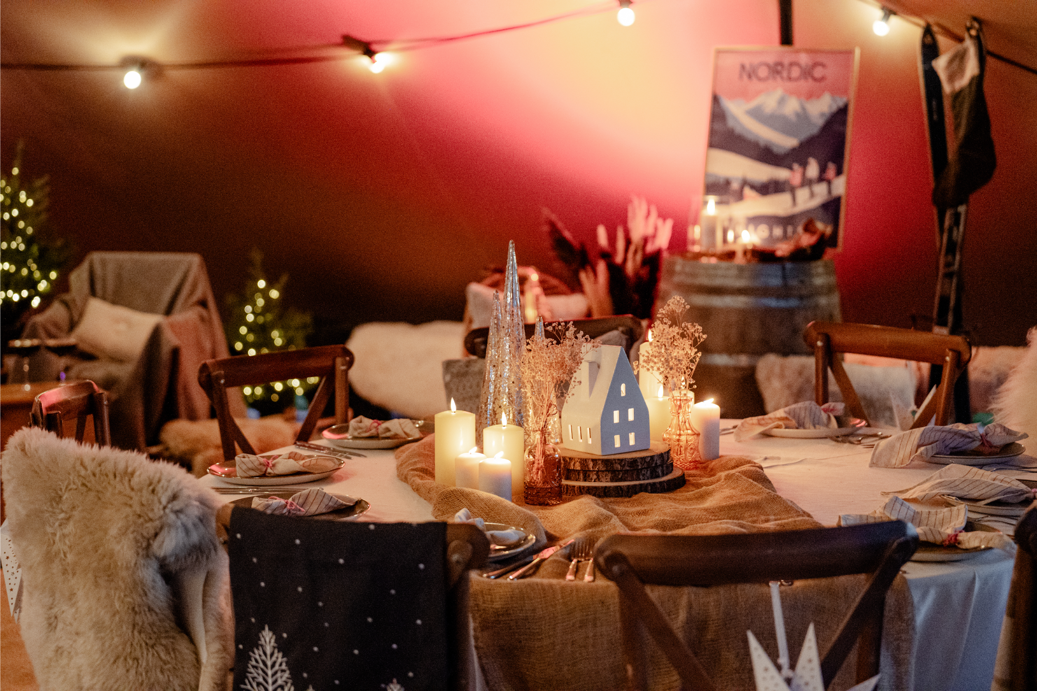 Decorated round dining table with candles, vases, and a small white house ornament, surrounded by wooden chairs with holiday and cozy decorations, in a warmly lit room with string lights.