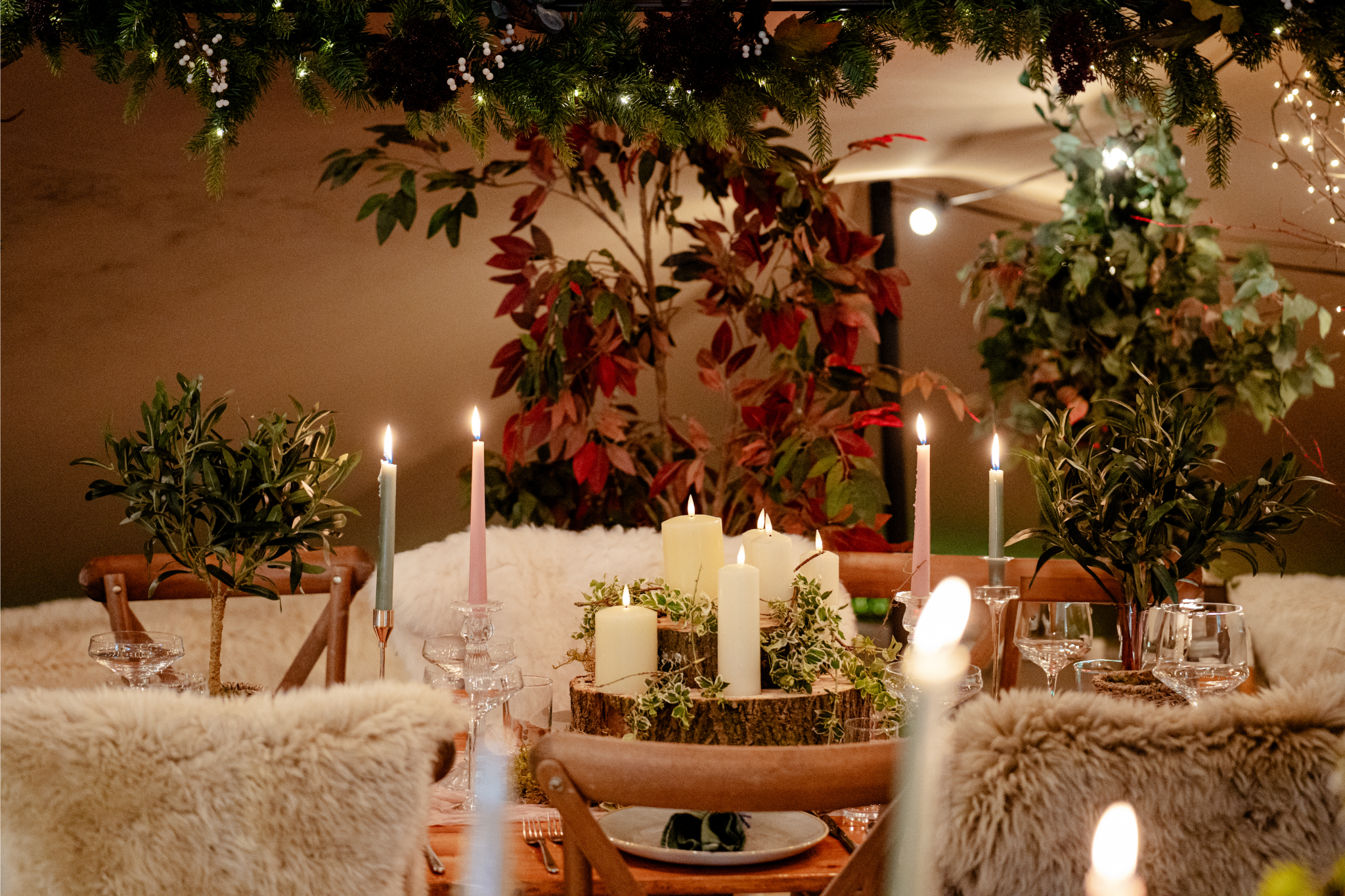 A festive dining table decorated with candles, greenery, and candles on a tree trunk centerpiece, surrounded by plush chairs and plants, with Christmas garland overhead.