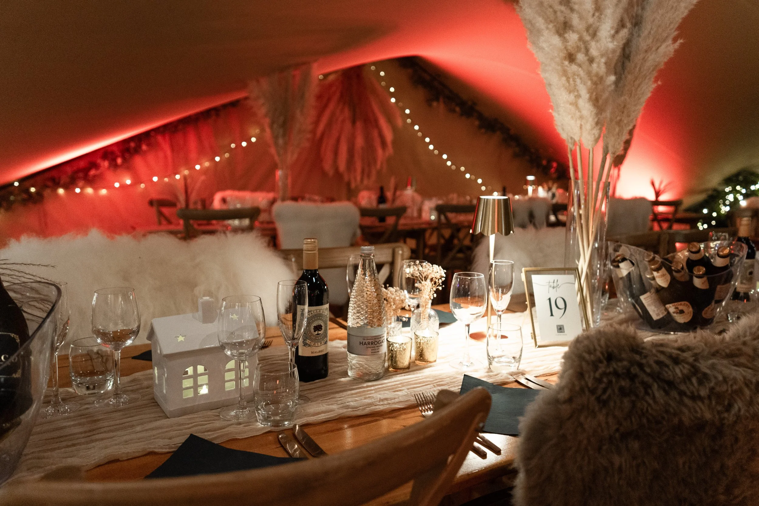 A decorated table set for a celebration in a dimly lit tent with pink and red lighting, featuring wine bottles, glasses, candles, a small house-shaped lantern, and a large vase with tall, fluffy pampas grass.