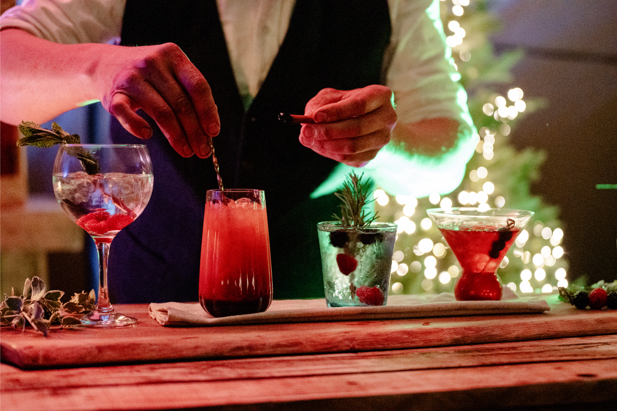 A bartender preparing holiday cocktails with garnishes including berries and herbs, set on a wooden bar with festive lights and a blurred Christmas tree in the background.