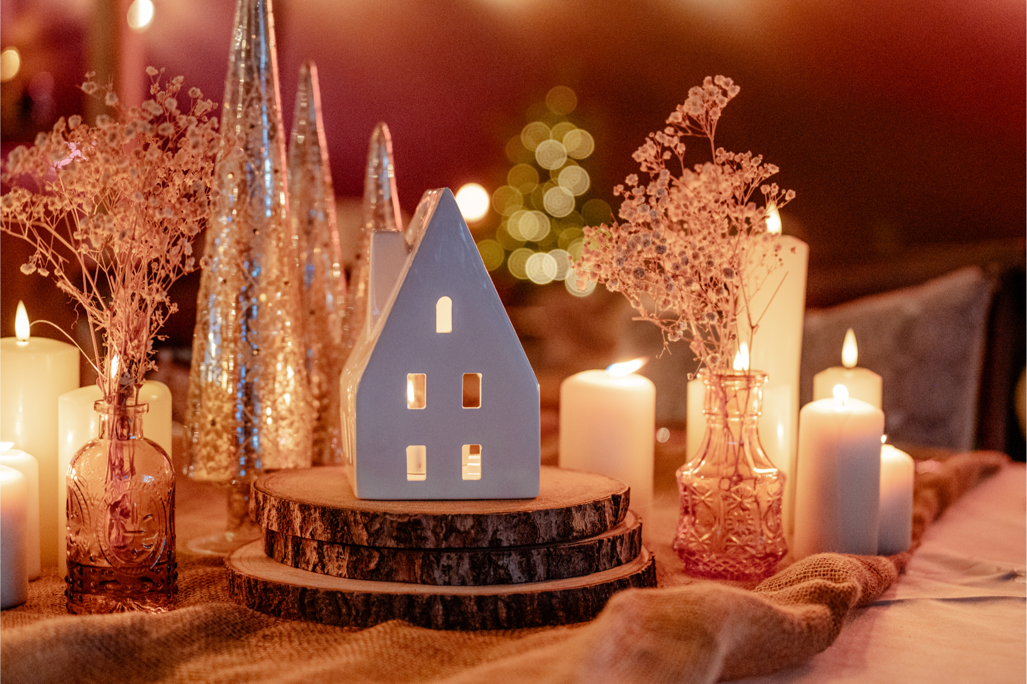 A cozy holiday table centerpiece featuring a small white ceramic house with cut-out windows, pink dried flowers in pink glass vases, and ivory candles on a wooden log slice, with blurred warm lights in the background.