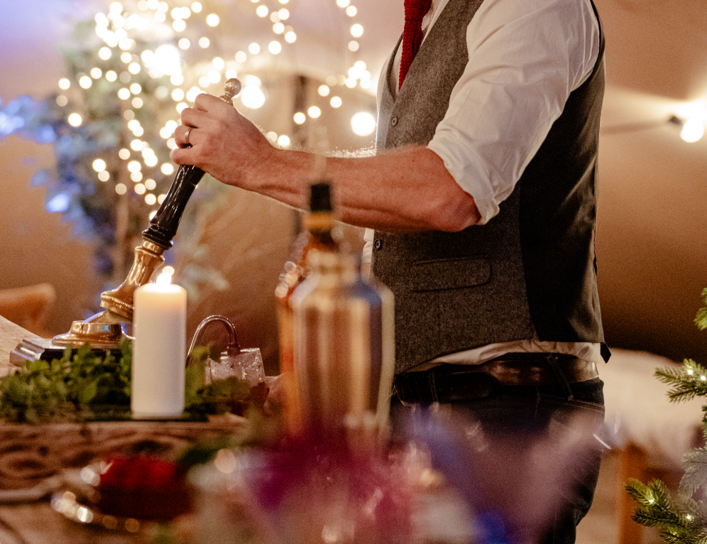 A person standing at a bar pouring a drink from a tap, with a lit candle and bottles on the table in front, and festive lights and decorations in the background.