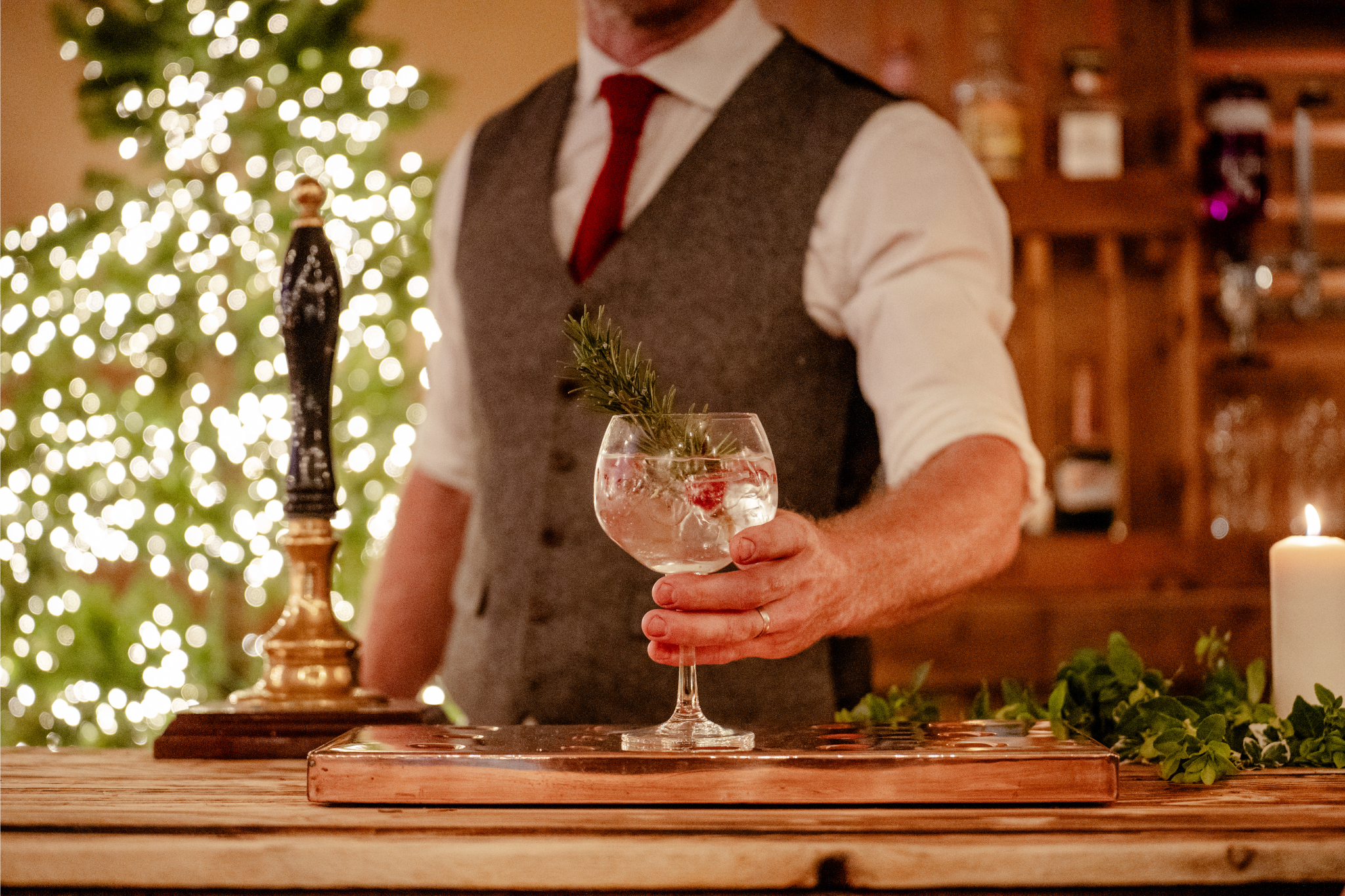 A man in a white shirt, red tie, and grey vest holding a glass with a sprig of rosemary and red berry inside, standing at a wooden bar with a Christmas tree and warm lighting in the background.