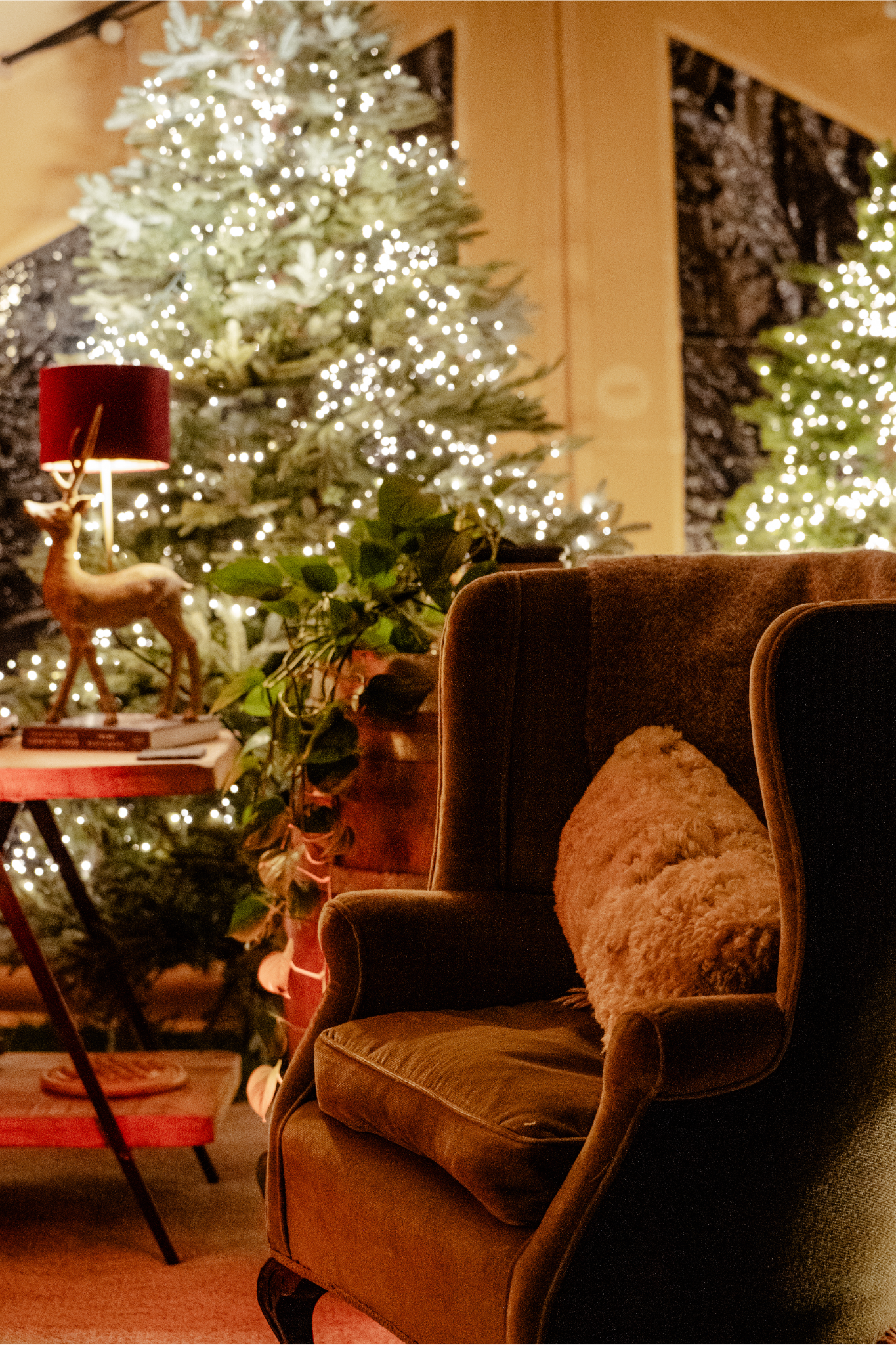 A cozy living room decorated for Christmas with a lit Christmas tree in the background, a plush armchair with a fluffy pillow, and a side table with a decorative reindeer figure and books, illuminated by warm lighting.