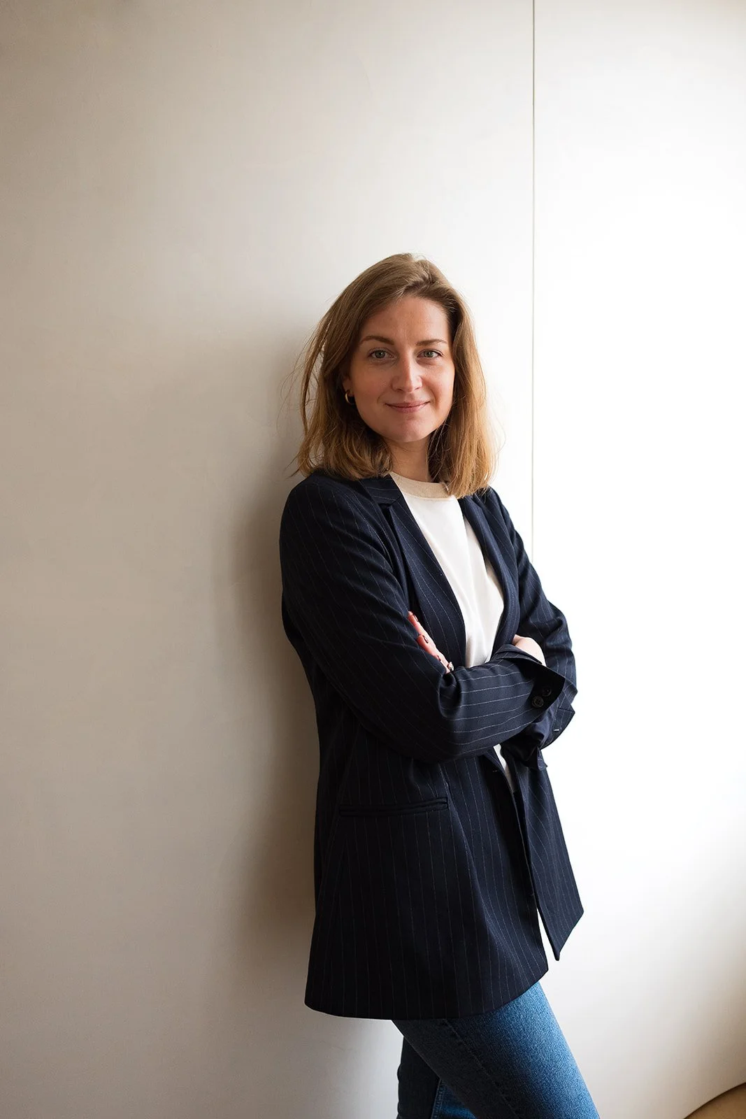 Jeune femme debout, professionnelle, posant contre un mur blanc, portant une veste blazer sombre et un t-shirt blanc.