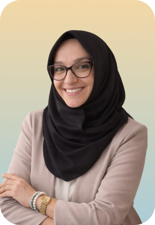 A woman smiling, wearing glasses, a black hijab, a beige blazer, and a white top, with a multicolored beaded bracelet and gold watch, standing against a light-colored background.