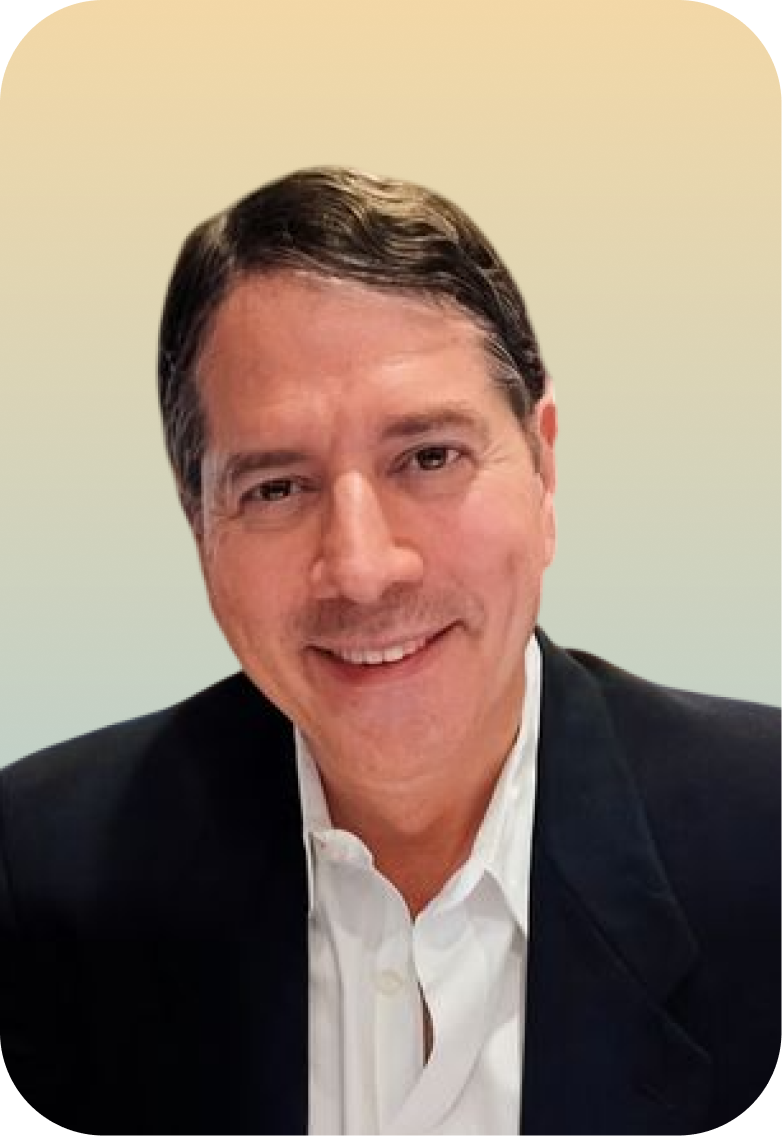 A man with dark hair, wearing a black blazer and a white shirt, smiling in front of a light-colored background.