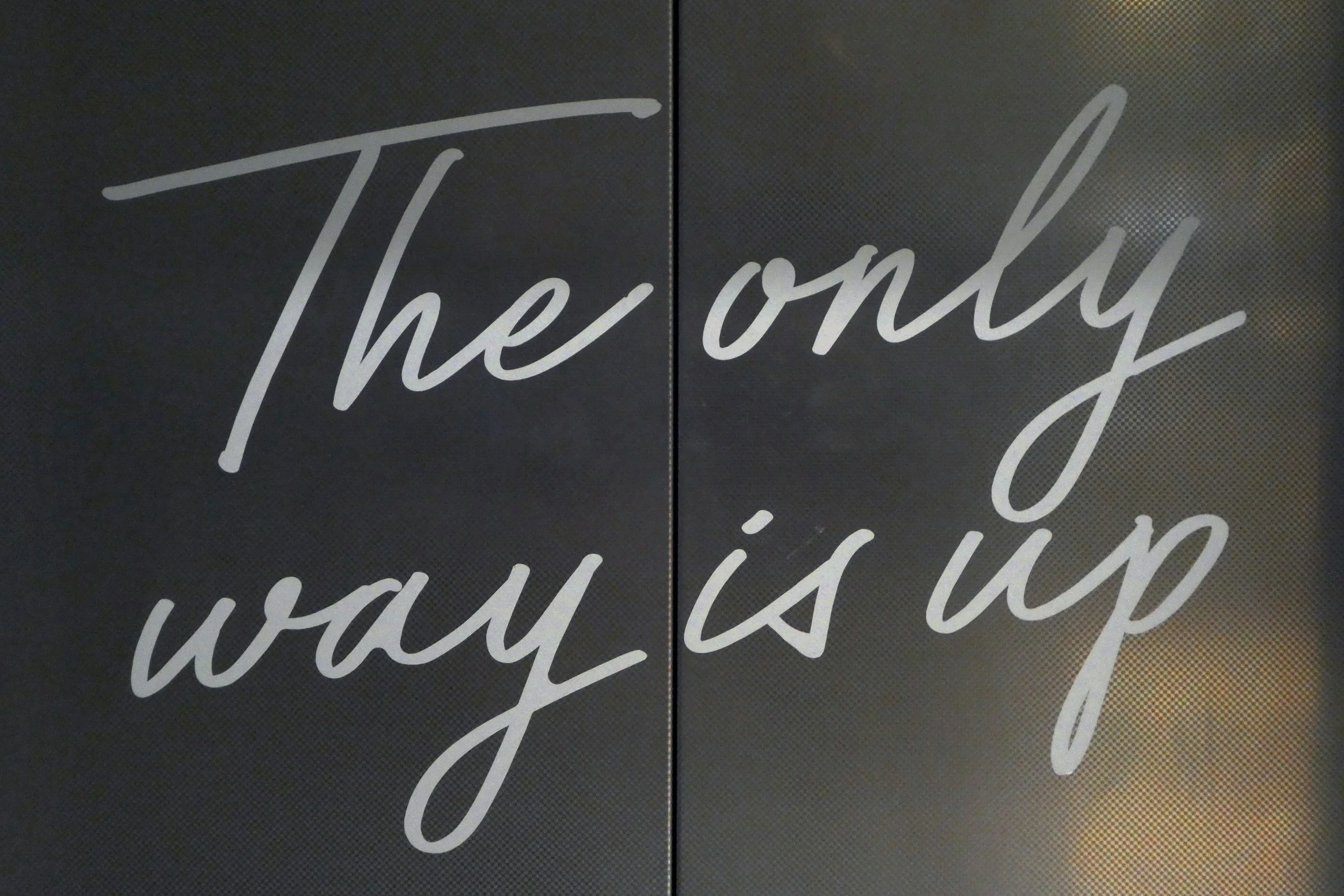 Close-up of a sign with the phrase 'The only way is up' in cursive writing on a dark background.