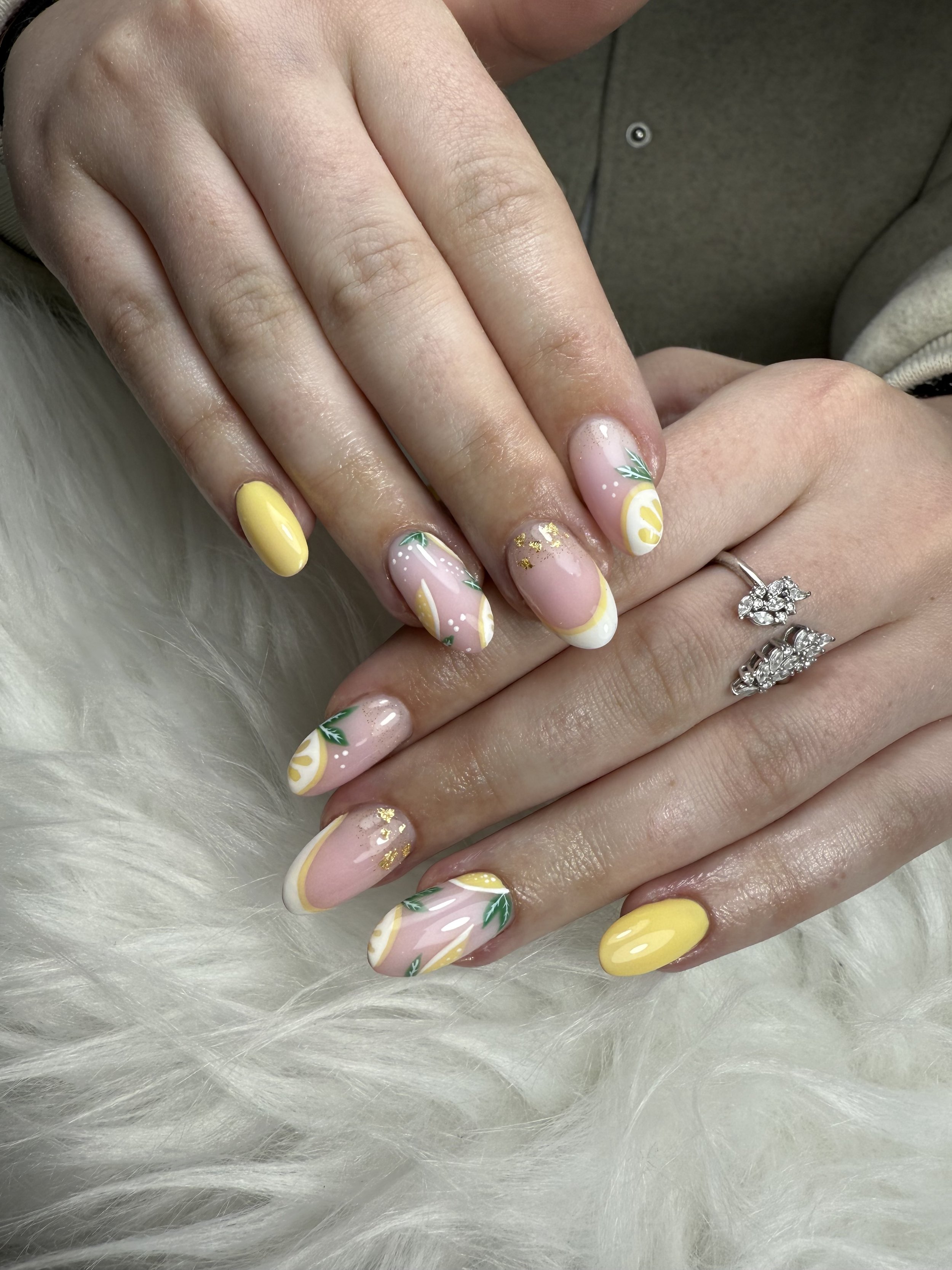 Mains avec des ongles décorés de vernis jaune et de motifs artistiques, incluant des feuilles et des touches dorées, sur fond de tissu blanc duveteux.