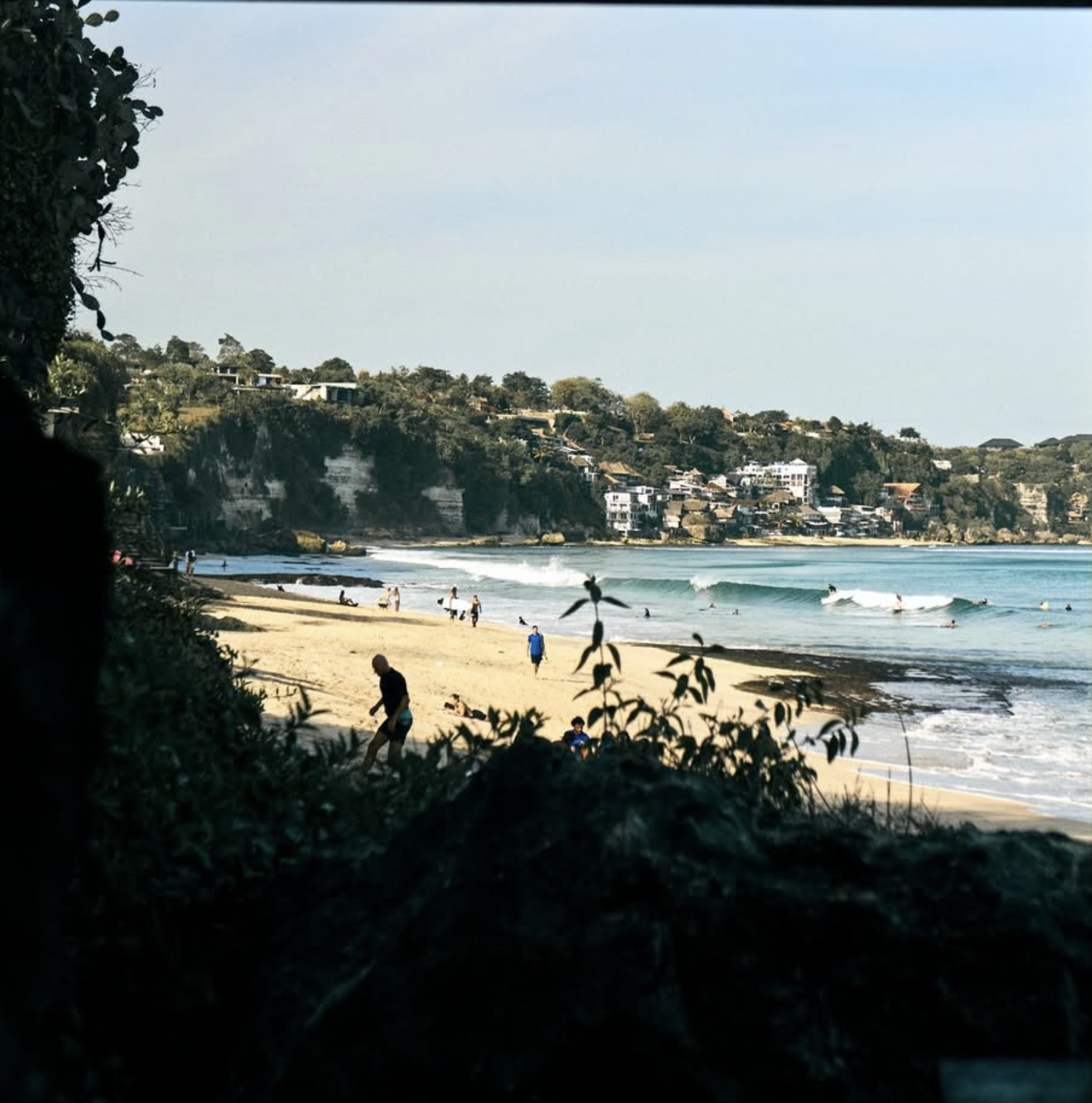 Olo Surf & Nature Balangan Bali. A beach scene with people walking and surfing, framed by foliage in the foreground, with houses on a hillside in the background under a clear blue sky.