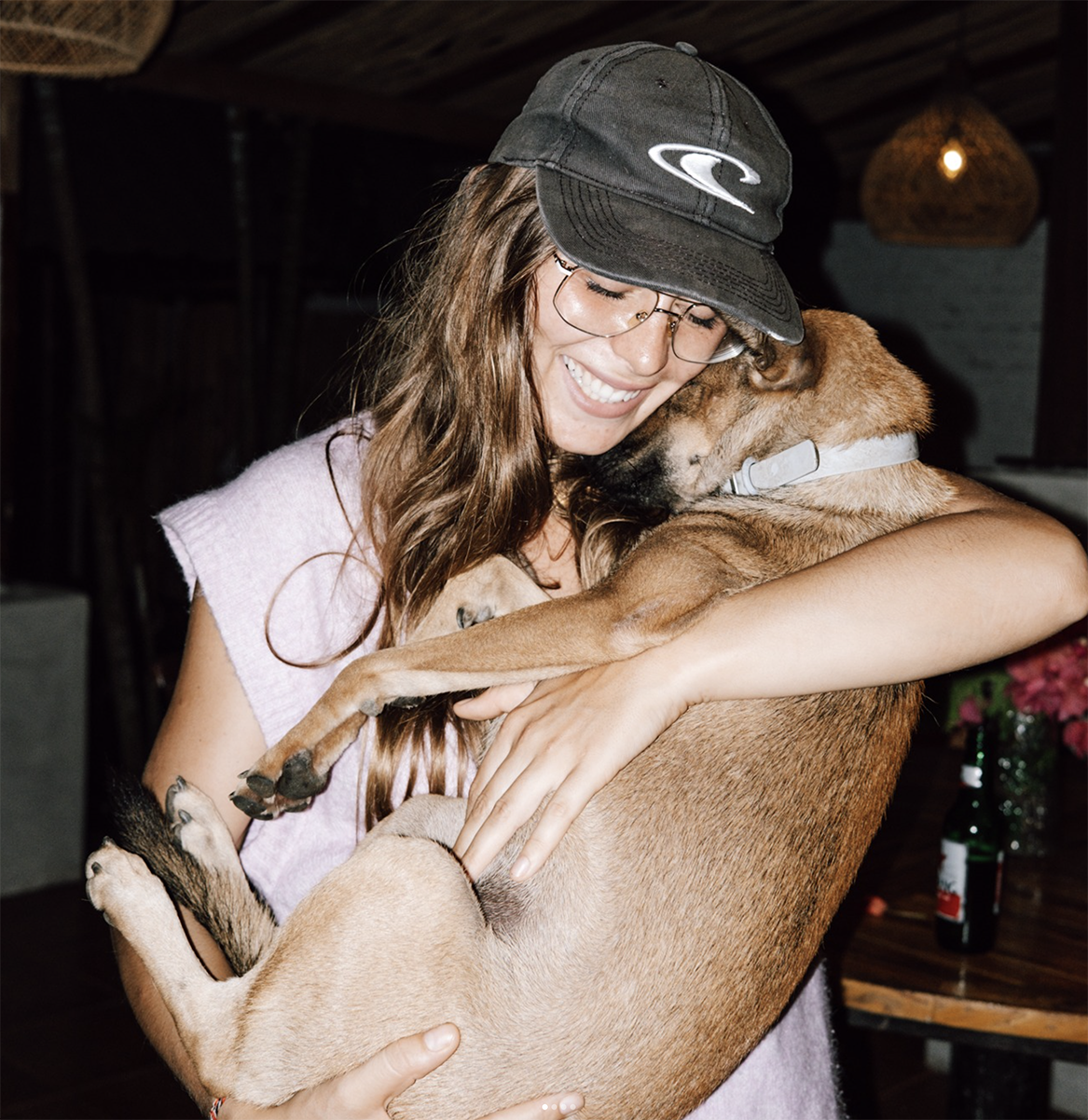 Olo surf & nature. A woman with glasses and a cap hugging a brown dog affectionately indoors.