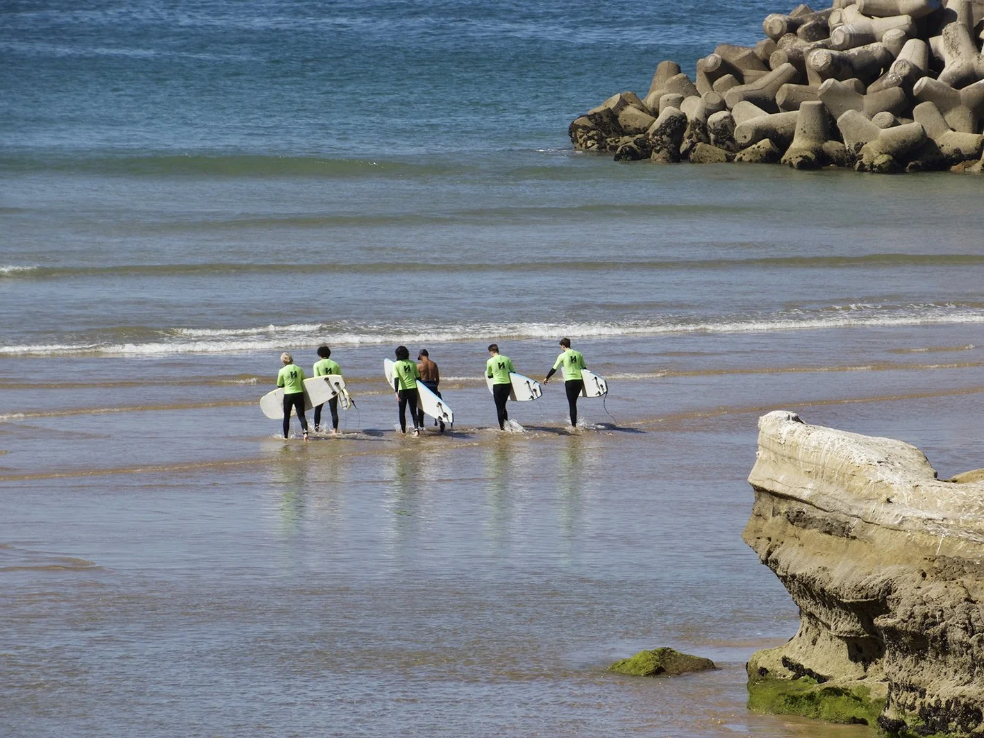 Olo surf & nature. Group of six surfers with surfboards and wetsuits standing in shallow ocean water near rocky shoreline.