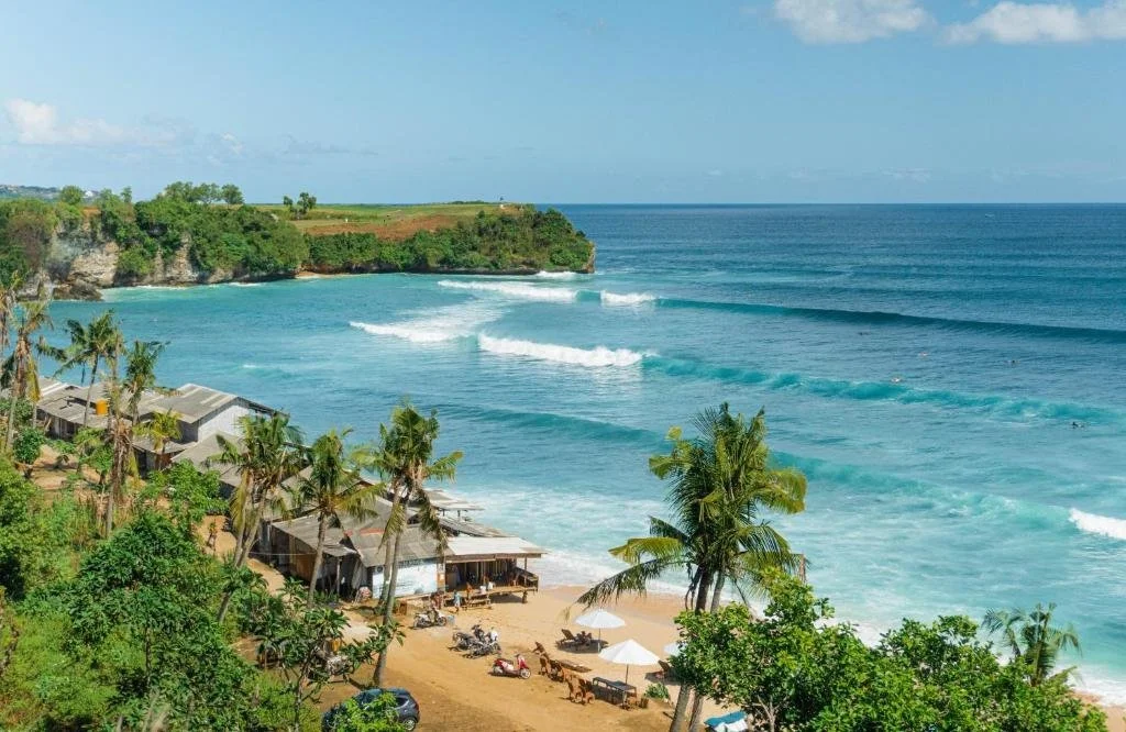Olo surf & nature. Beach with turquoise water, palm trees, small houses, and a green cliff in the background under a partly cloudy sky.