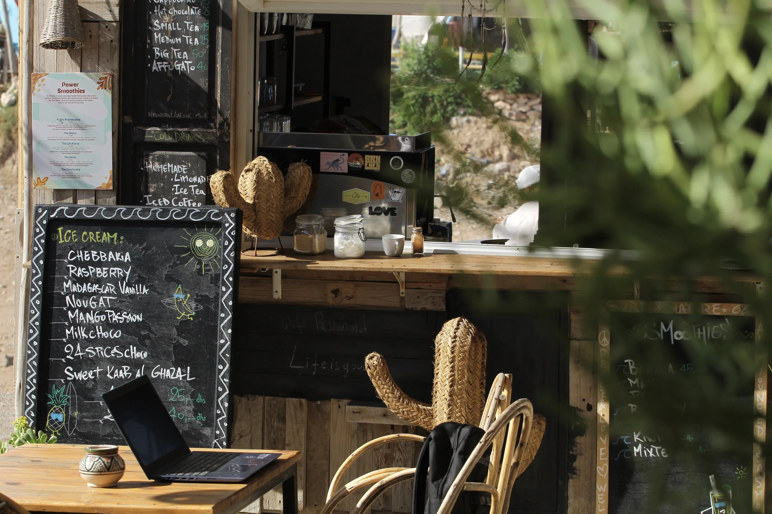 A rustic outdoor cafe stall with a black chalkboard menu displaying ice cream flavors, a laptop on a wooden table, decorative woven hats, a pineapple drawing on the chalkboard, and a partly blurred background with greenery.