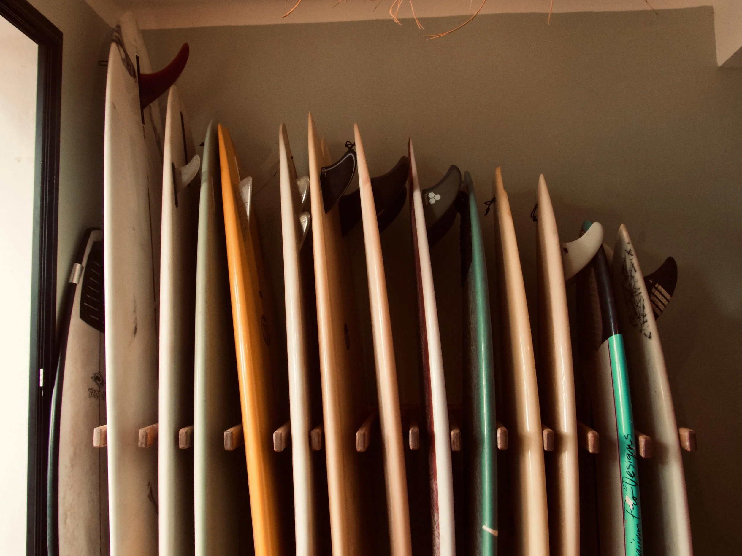 Olo surf & nature. Multiple surfboards of various colors and sizes standing upright against the wall near a window.