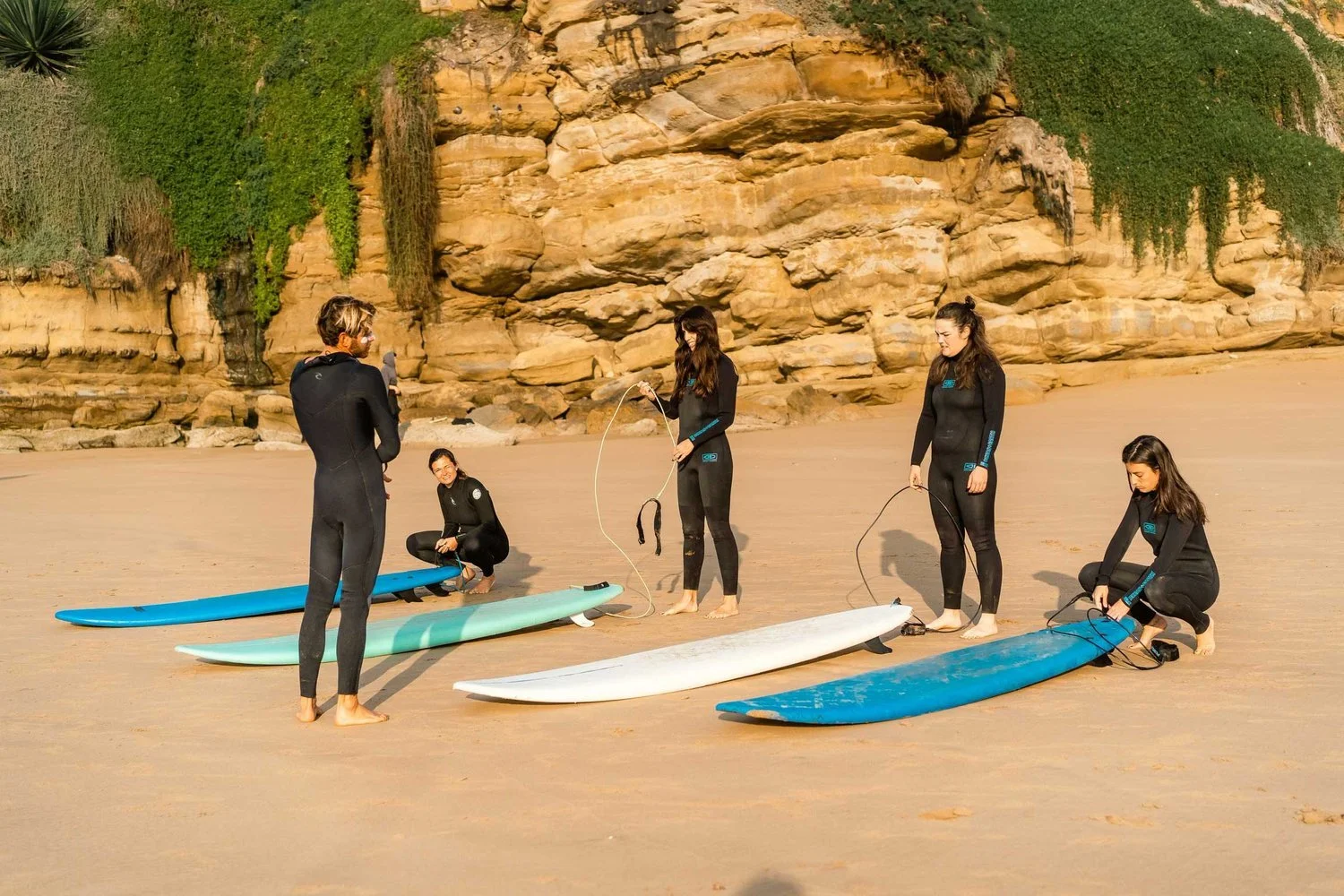 Olo surf & nature. Four women in wetsuits preparing surfboards on a sandy beach with rocky cliffs and greenery in the background, engaging in a surfing instruction or lesson.