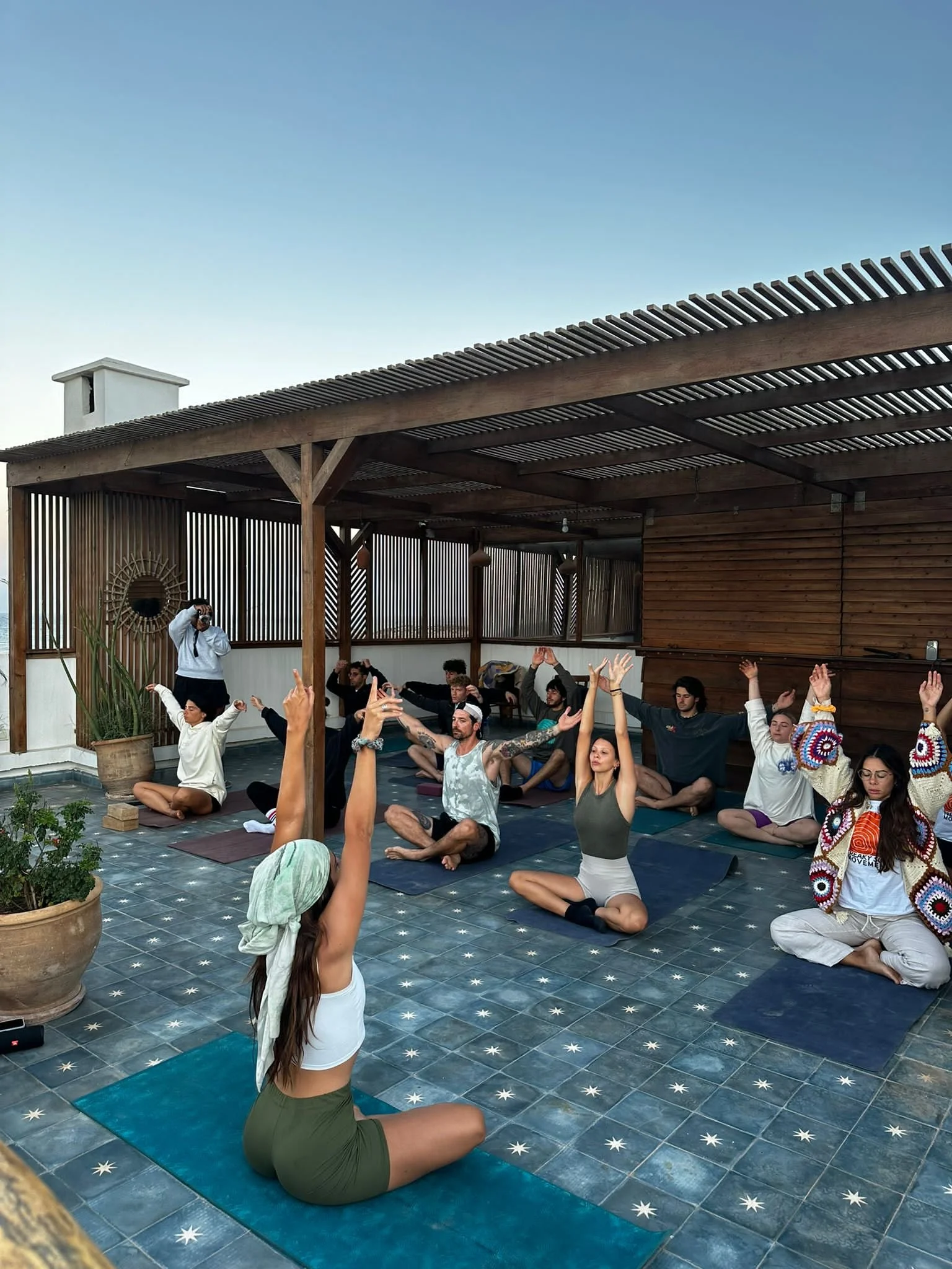 Group of people practicing yoga on a rooftop terrace during sunset, seated on yoga mats with arms raised, with a clear sky overhead.