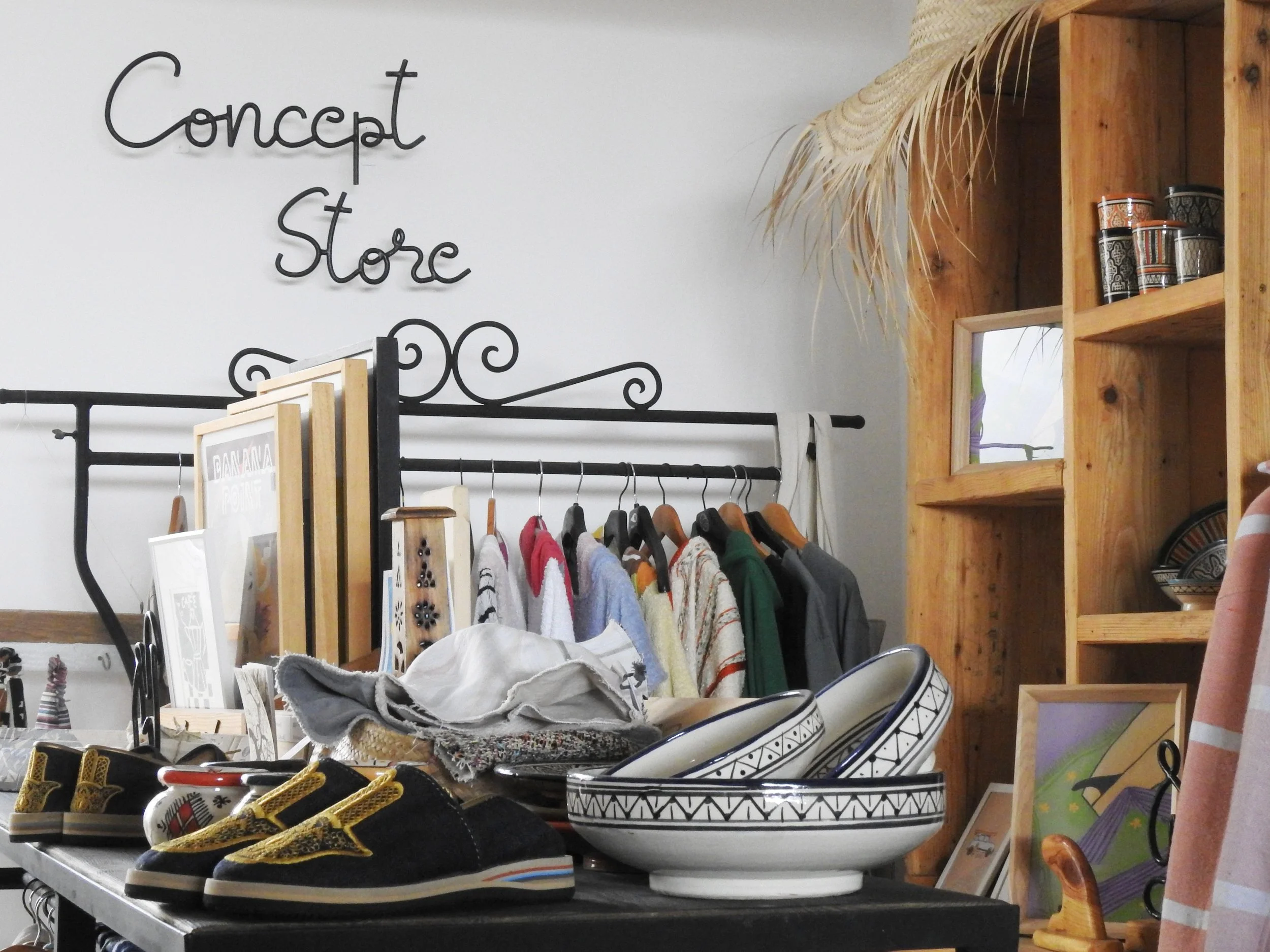 Interior of a boutique store with clothes on a rack, shoes, bowls, and decorative items on tables and shelves, and a sign that says 'Concept Store' on the wall.