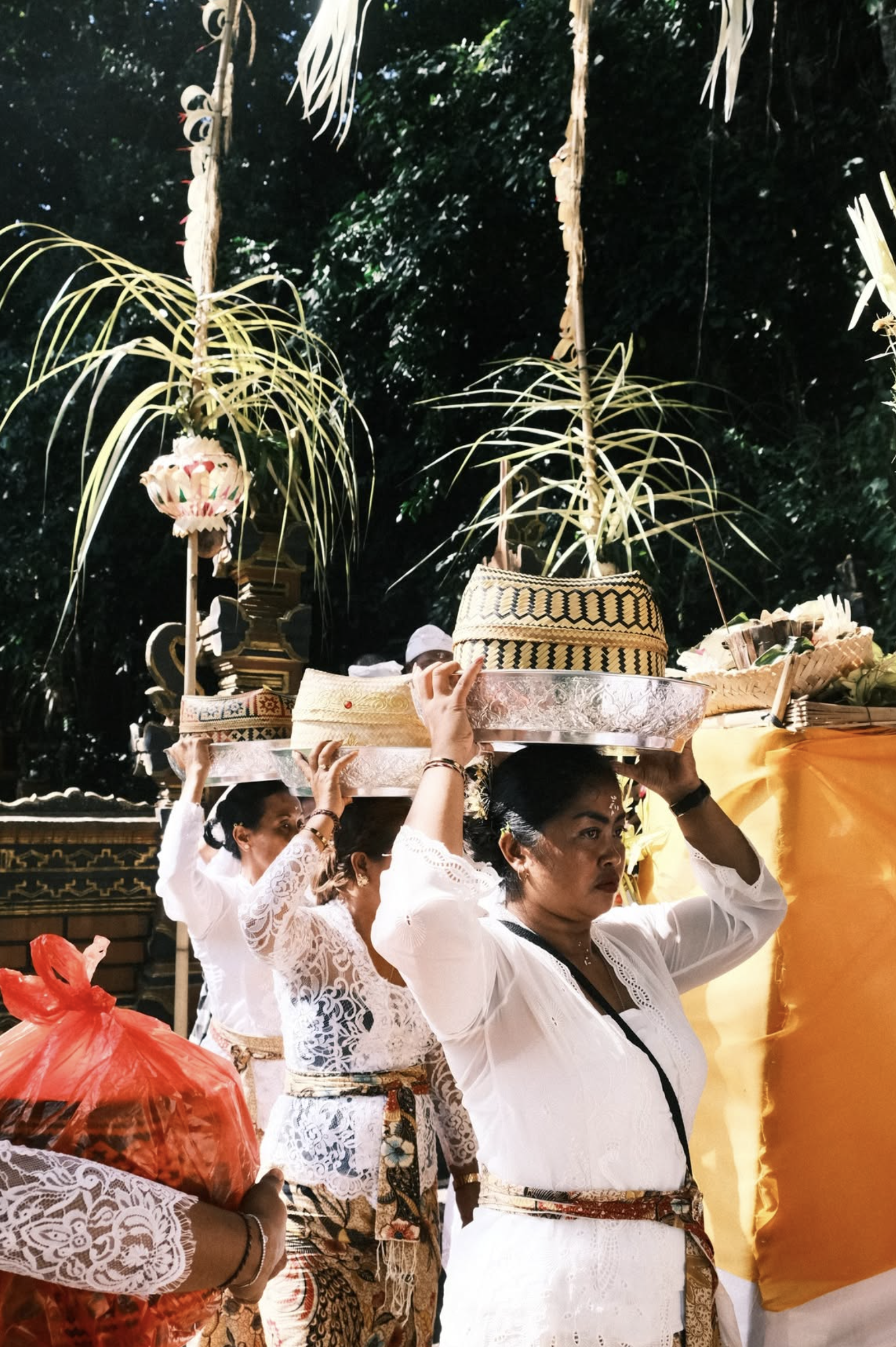 Olo surf & nature. Women dressed in traditional white dance attire carrying decorated trays with offerings on their heads during a cultural or religious ceremony outdoors, with green foliage in the background.