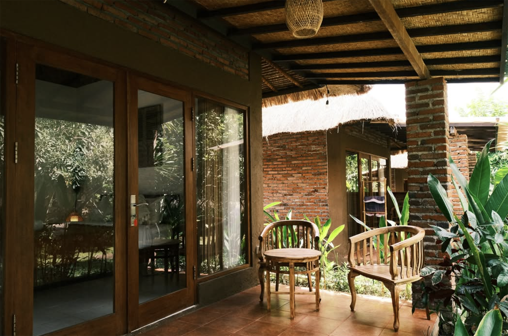 Olo surf & nature. Olo surf & nature. A rustic outdoor patio with wooden furniture, brick walls, glass sliding doors, green plants, and a thatched roof.