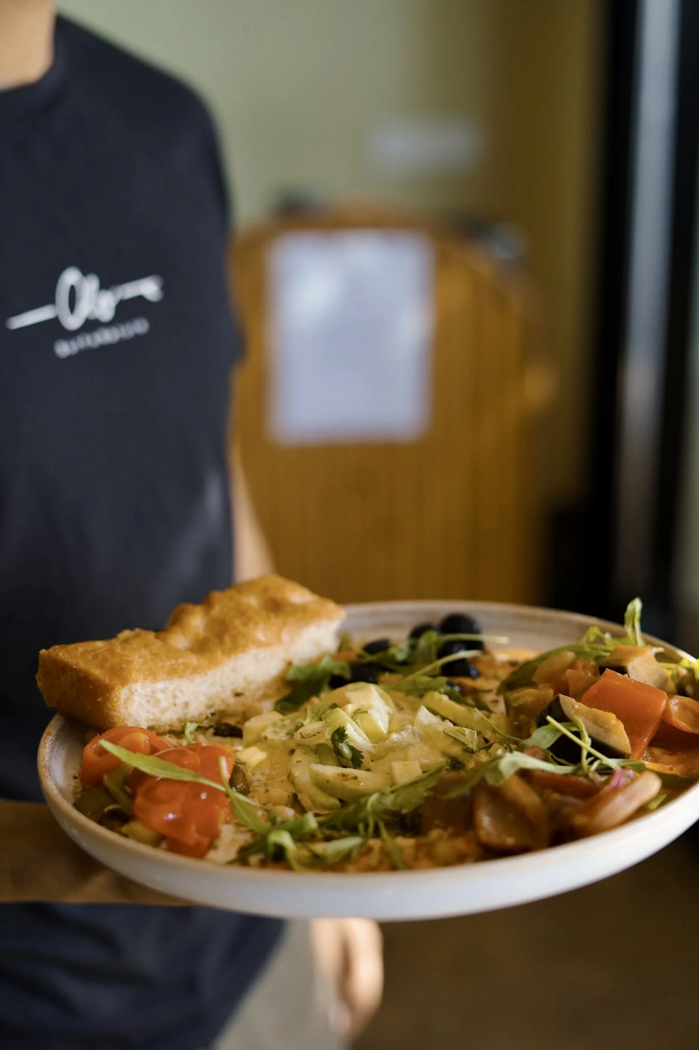 Person holding a white bowl of assorted vegetarian or vegan food, topped with a slice of bread, inside a restaurant or cafe.