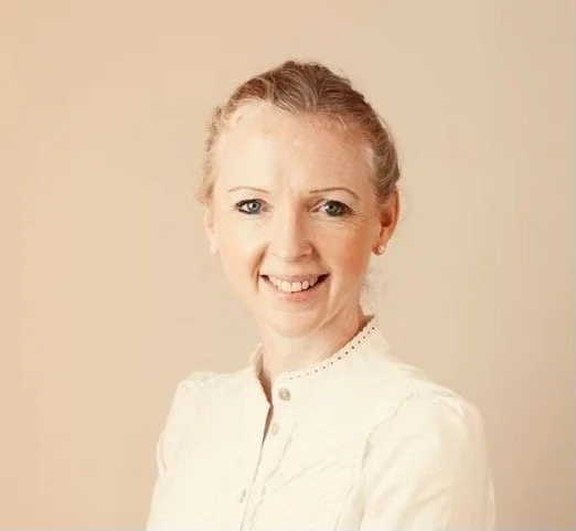 A woman with light skin, blonde hair, wearing a white blouse, smiling, against a beige background.