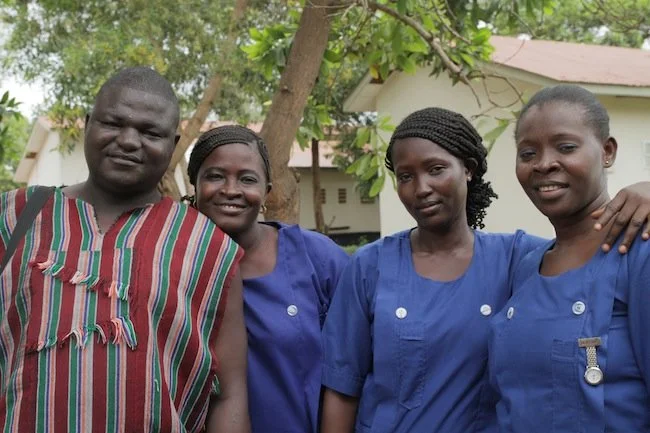 Four people standing outdoors near a tree, three women wearing blue uniforms and one man in traditional colorful clothing, smiling.