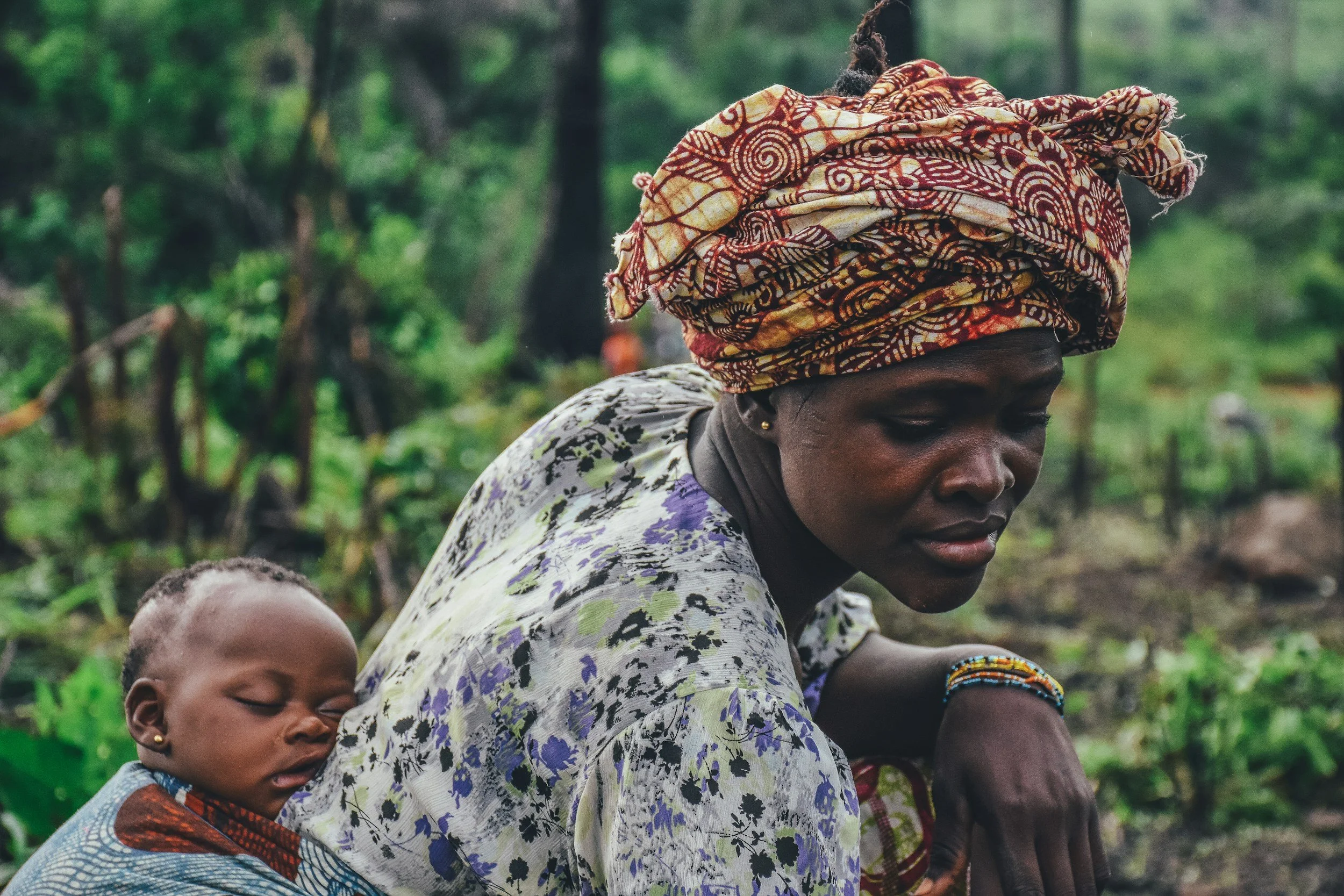 A woman wearing a colourful headscarf and patterned clothing with a sleeping baby attached to her back in a lush green outdoor setting.
