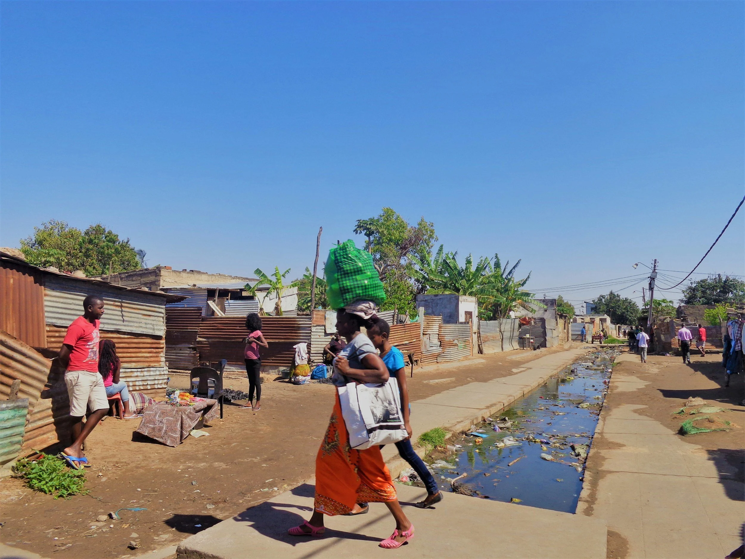A dirt street in a poor neighborhood with people walking and talking. Some are carrying bags, and a woman is balancing a large green bundle on her head. There are makeshift houses made of corrugated metal, a small canal filled with water and garbage, and utility poles with wires. The sky is clear and blue.